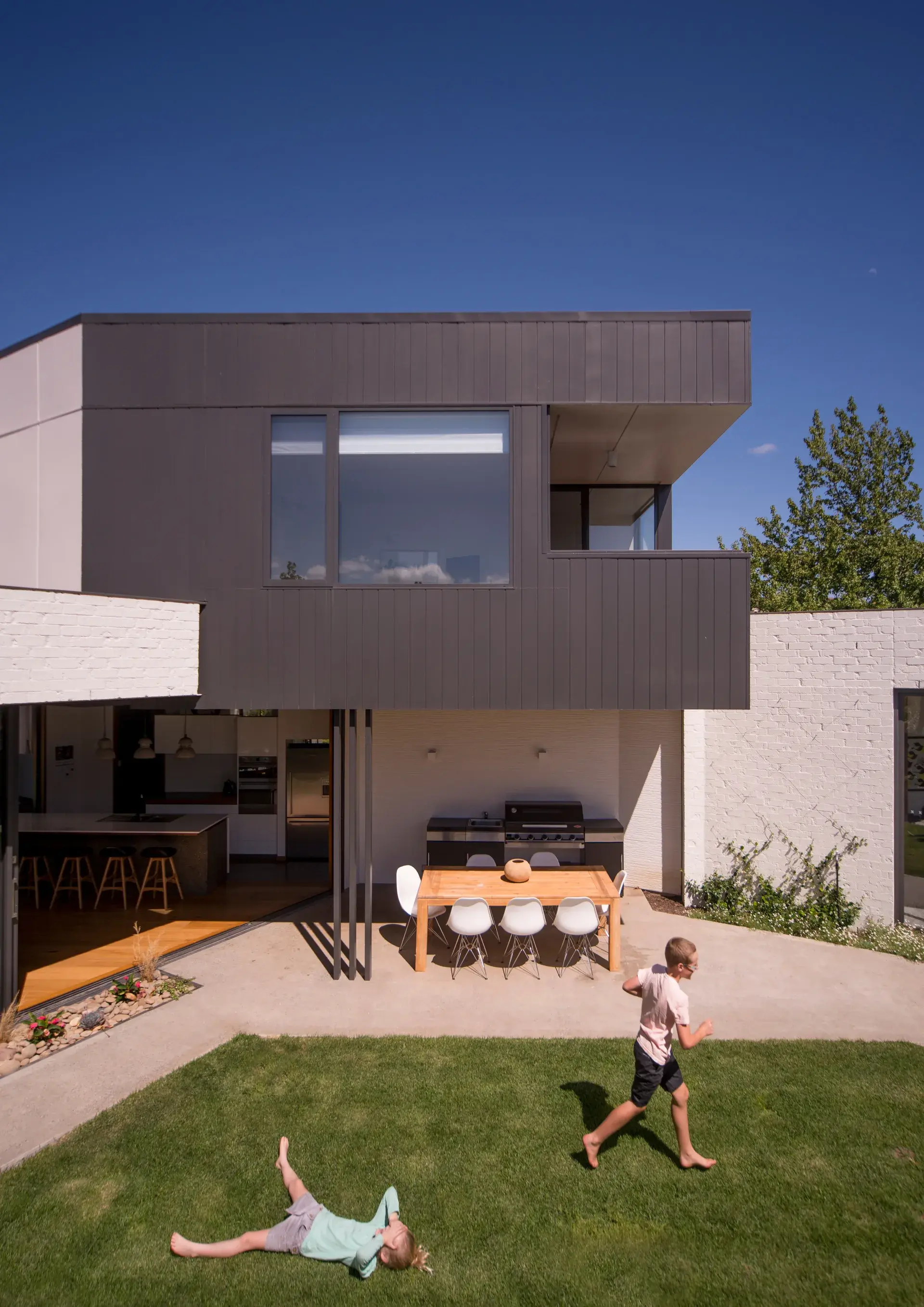 An exterior view of the upper-level volume expressed in dark vertical cladding, projecting over the courtyard to provide shade and enclosure, with a deep-set window and recessed balcony overlooking the outdoor living spaces with children playing.