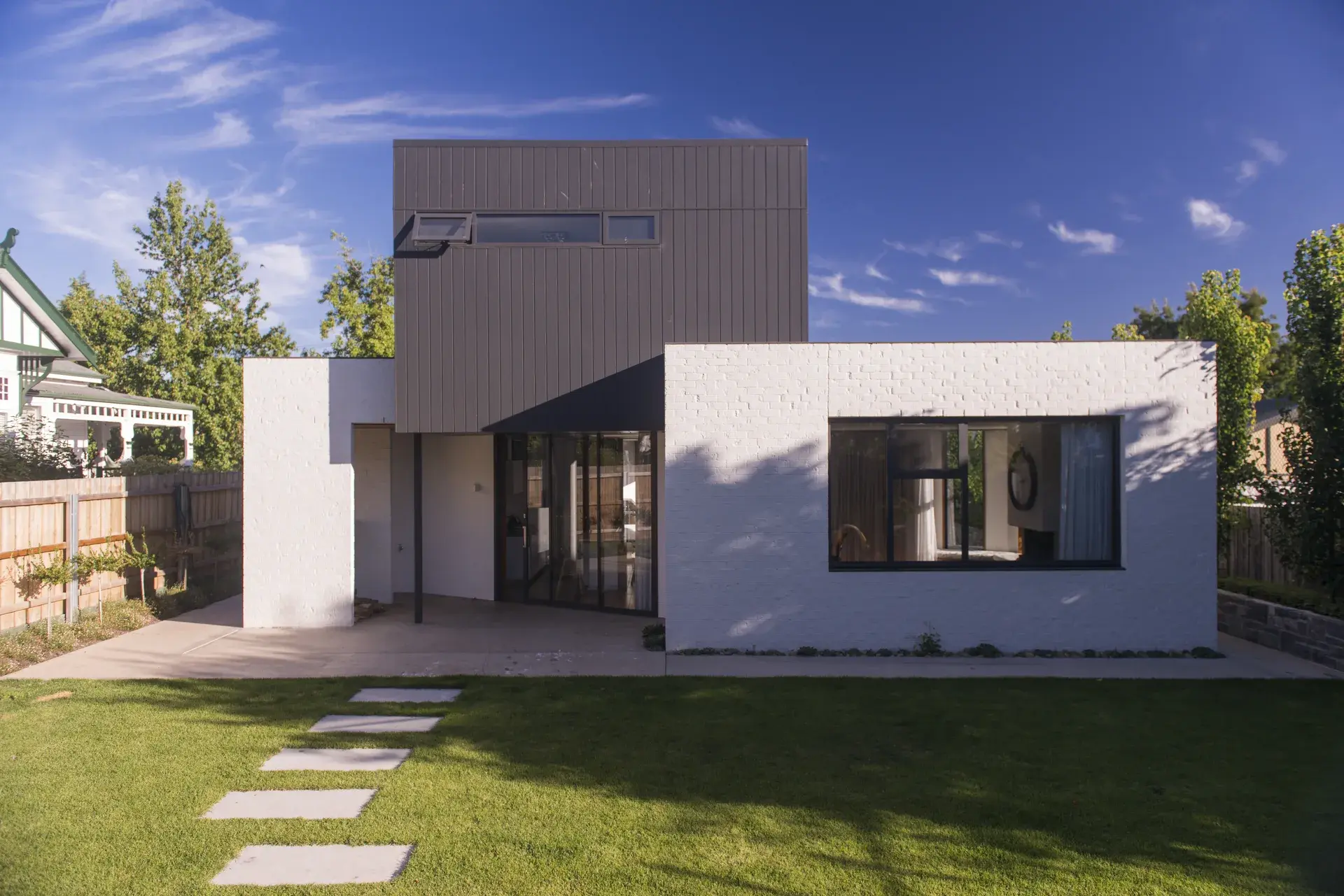 A photograph of the rear elevation showing the house opening to a central courtyard, with white-painted brick volumes at ground level and a dark, vertically clad upper form hovering above. Full-height sliding glazing connects living spaces to a shaded outdoor dining area and lawn.
