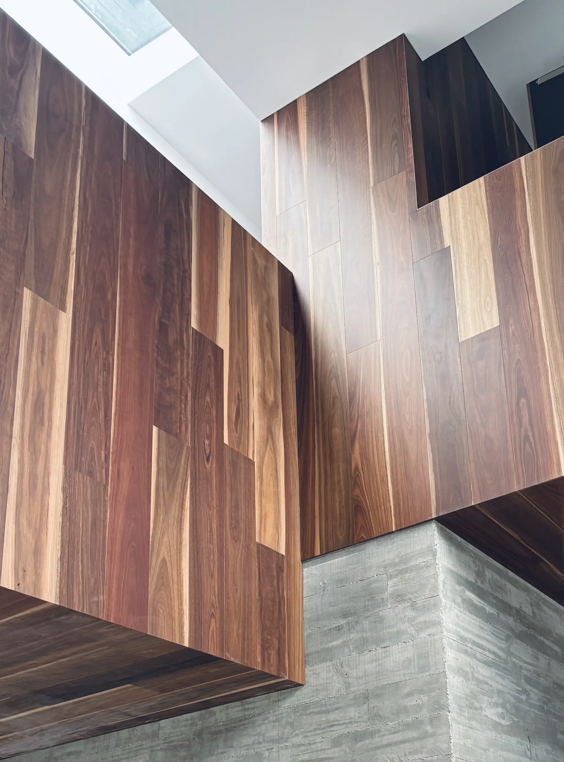 Detail of rich timber wall panelling meeting a raw concrete base, with a skylight casting natural light across the grain.