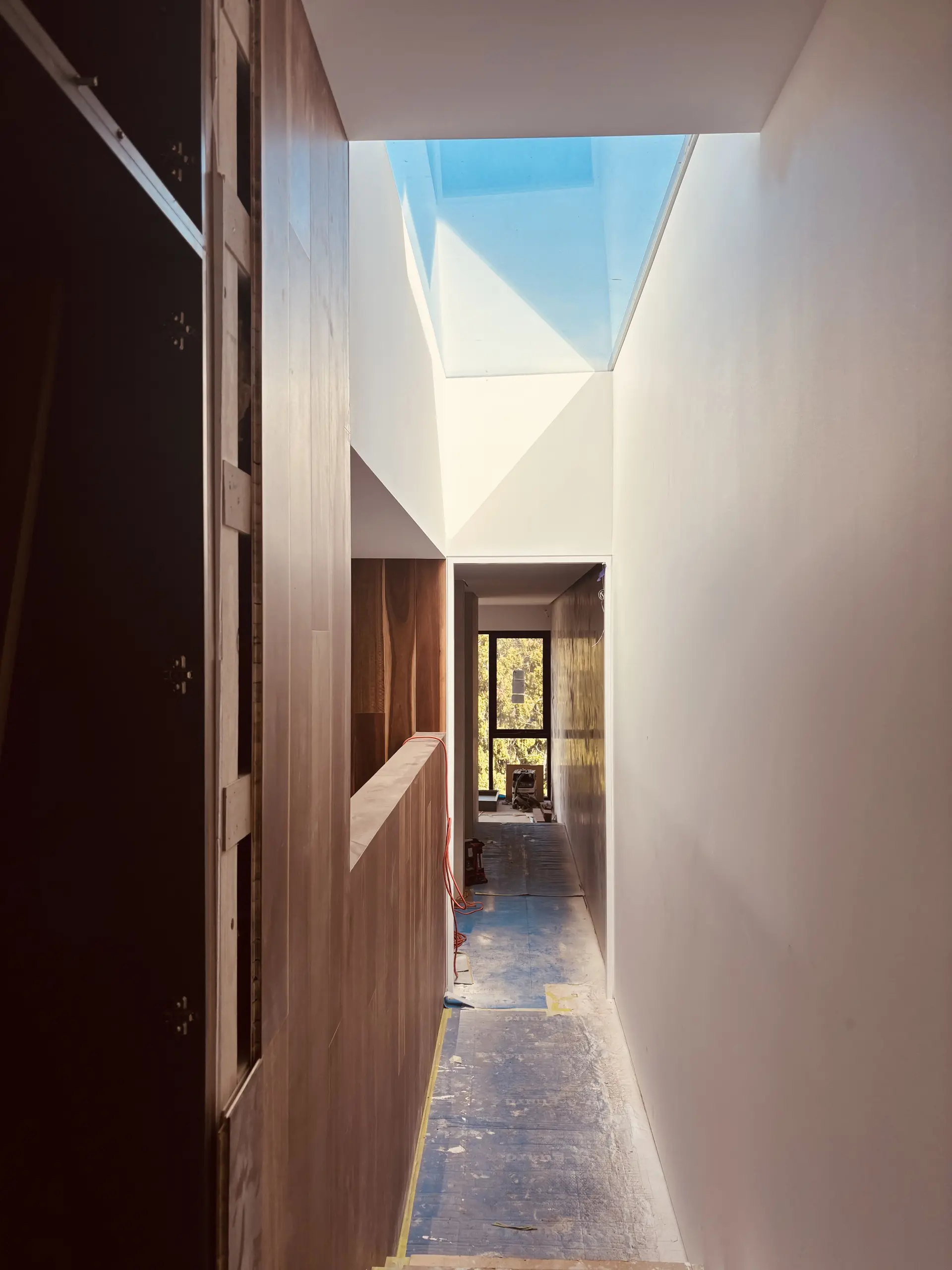 Interior corridor under construction, with timber joinery lining one wall, a skylight above, and views to tree canopy through a window at the end of the hall.