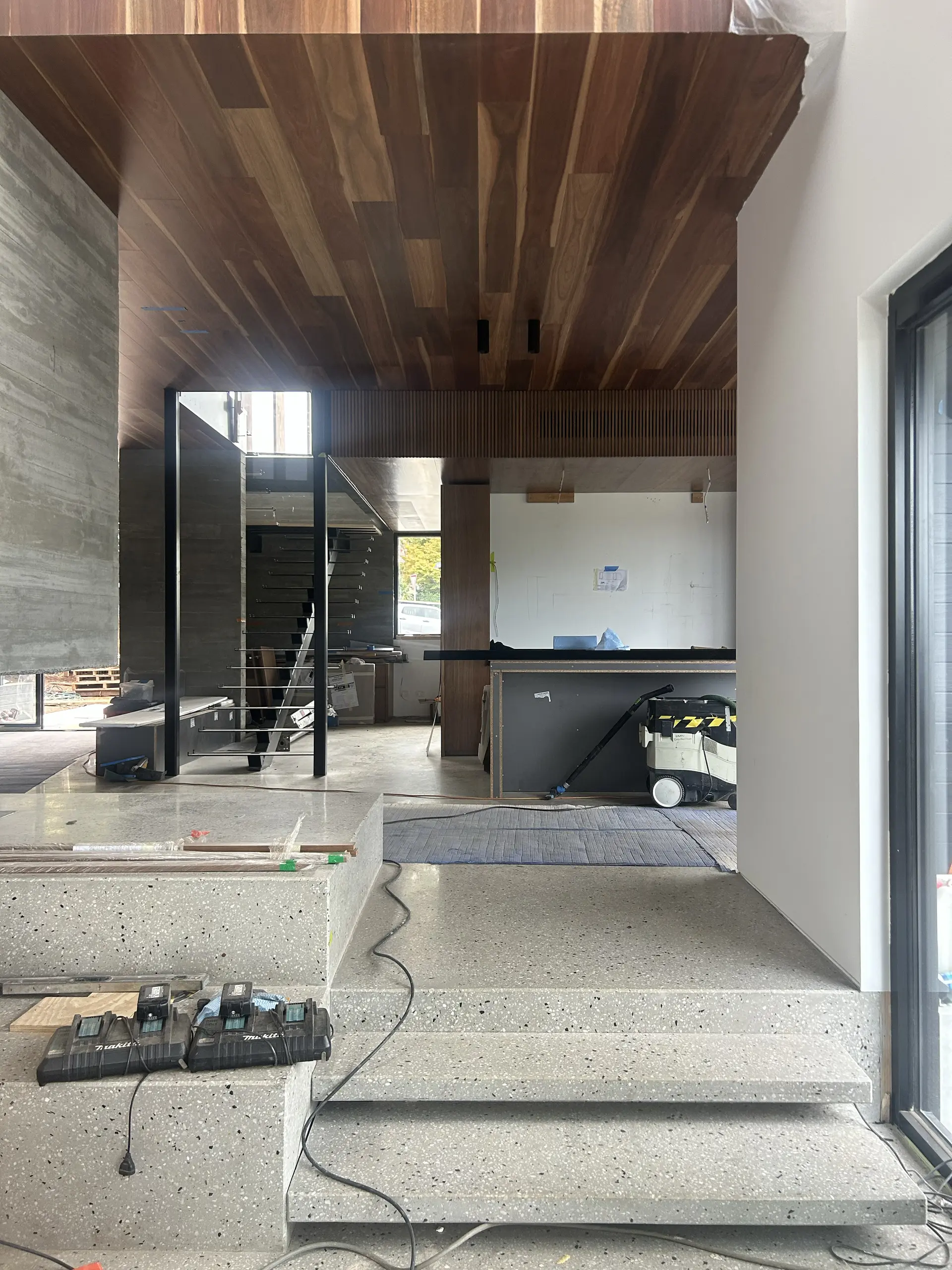 Interior under construction, showing split-level polished concrete steps in the foreground, a timber-lined ceiling, and kitchen joinery being installed beyond.