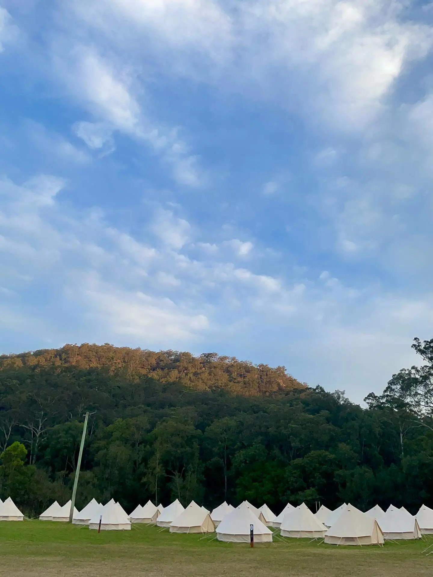 A row of white bell tents set up on a grassy field at the base of a densely forested hill under a blue, cloudy sky.