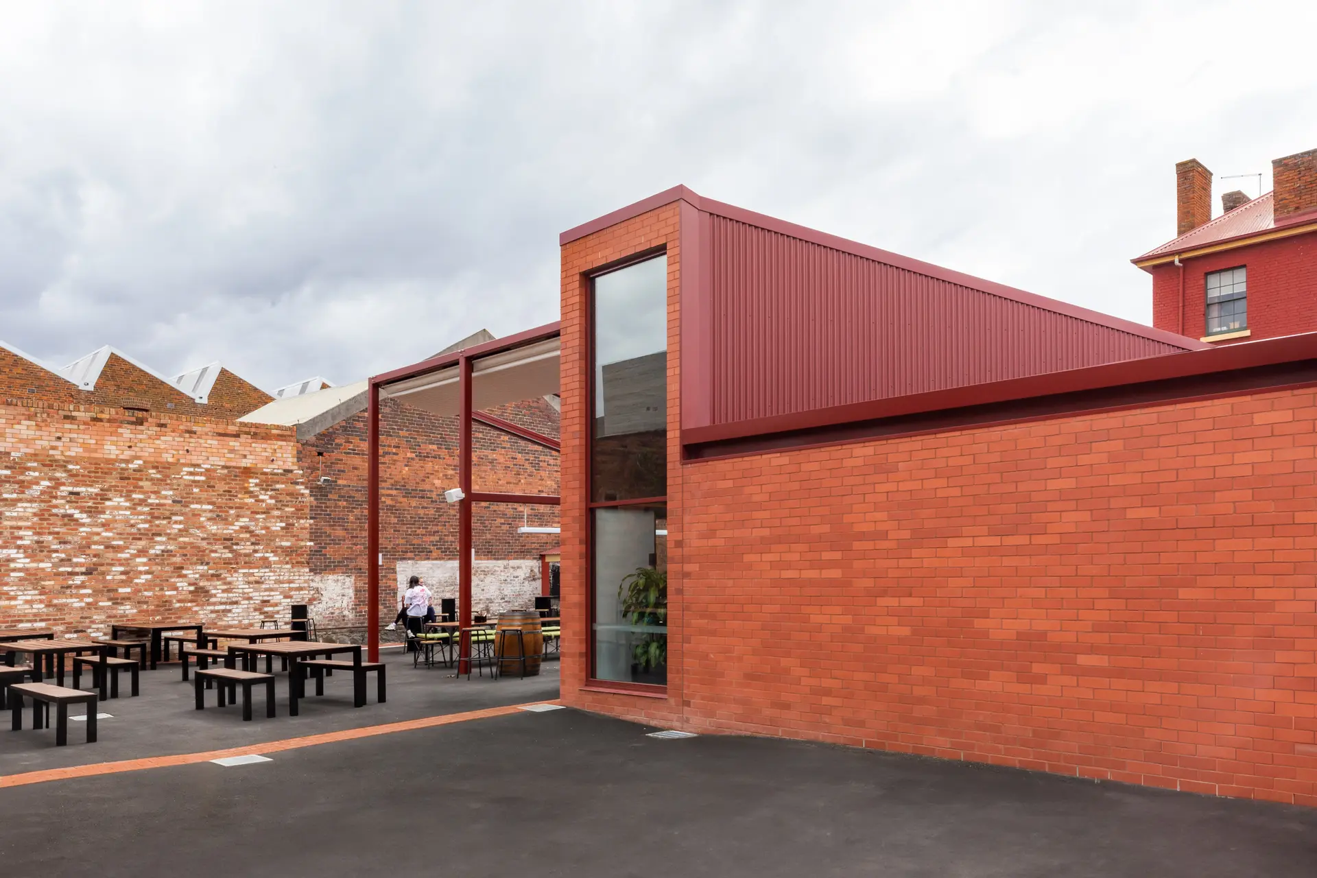 An outdoor courtyard with dark picnic tables next to a modern red brick and corrugated metal building extension, featuring an industrial urban backdrop.