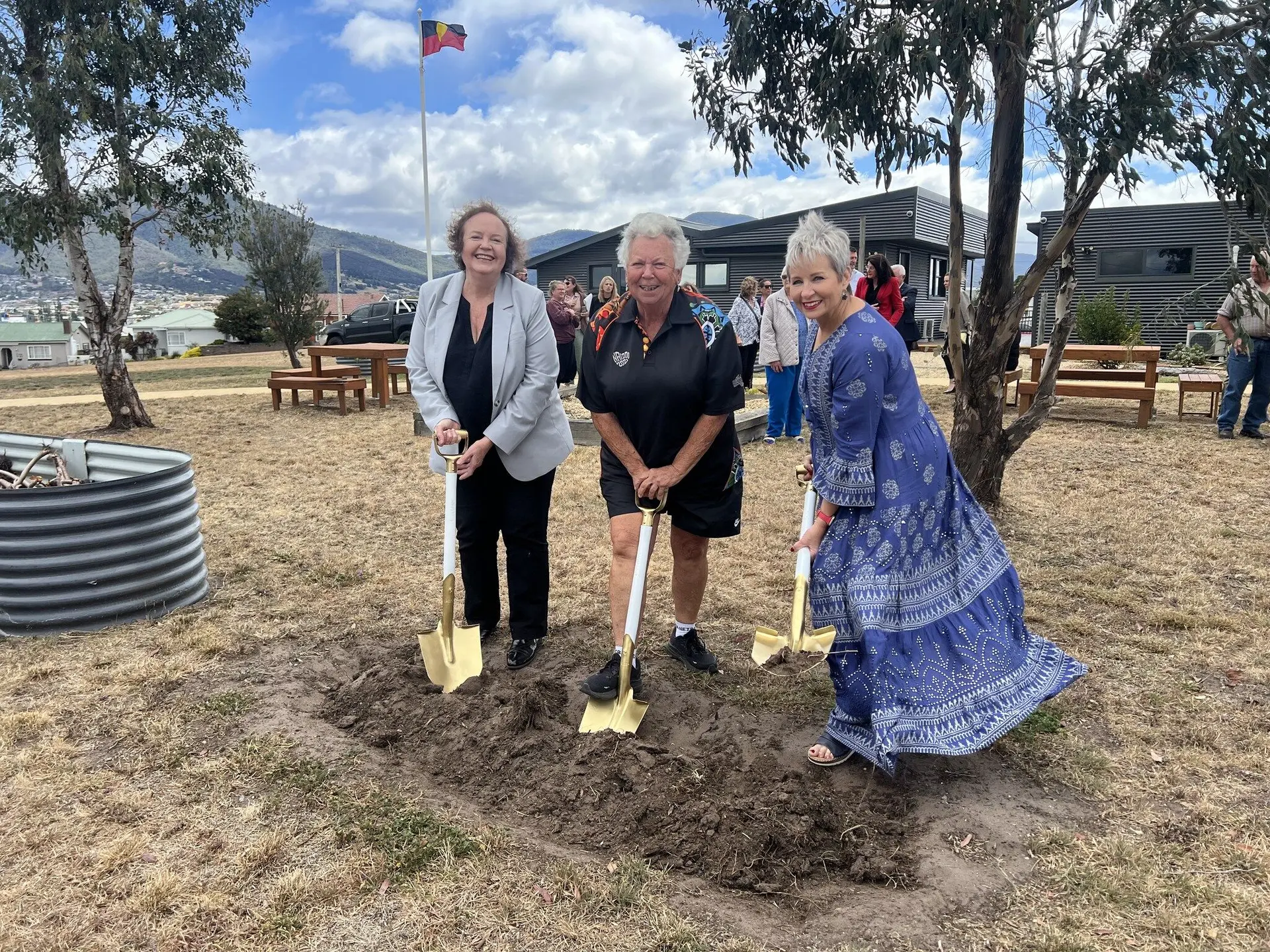 Three women smiling during a groundbreaking ceremony, holding gold-coloured shovels and turning soil in a grassy field with buildings and hills in the background.