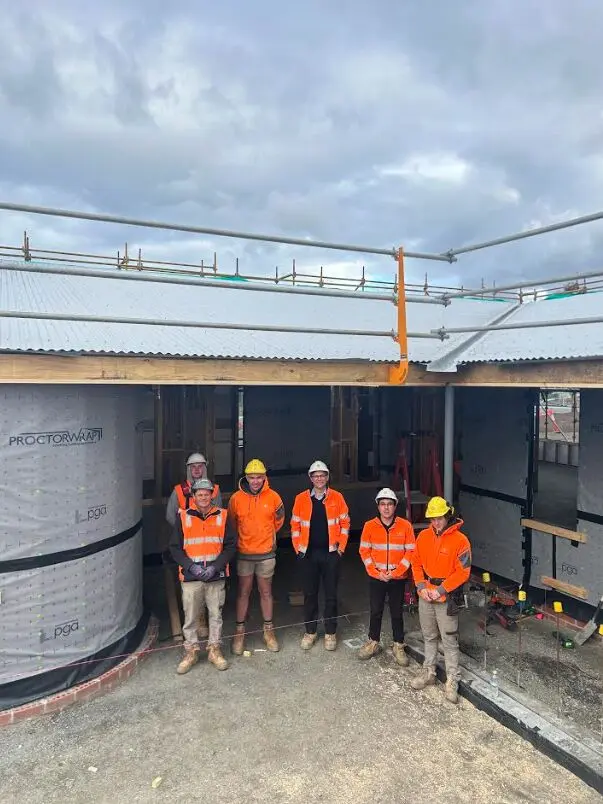 A group of six construction workers and professionals wearing orange high-visibility jackets and hard hats, posing together on a construction site in front of a building with visible insulation and roofing.