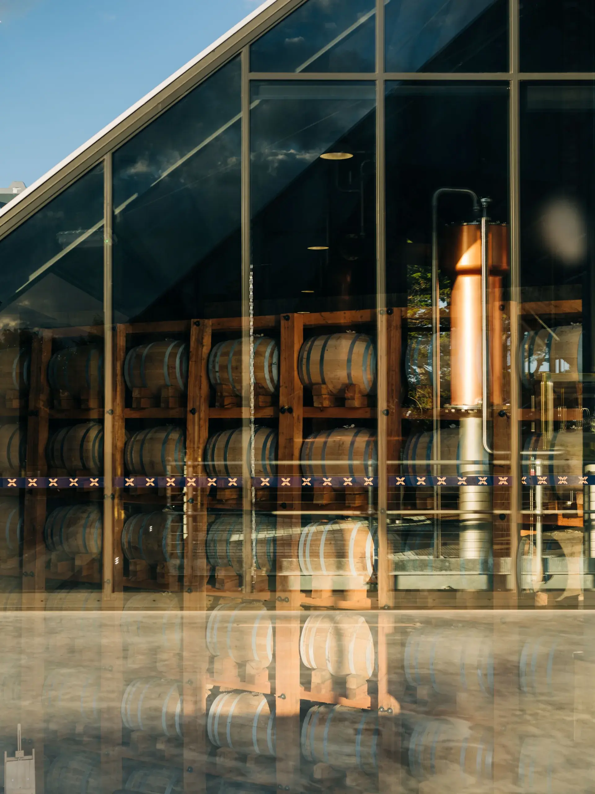 A close-up through a glass wall of a Tasmanian whisky distillery, showing rows of oak barrels stacked on wooden racks alongside a tall, gleaming copper pot still, with reflections of the modern architectural facade visible on the glass surface.
