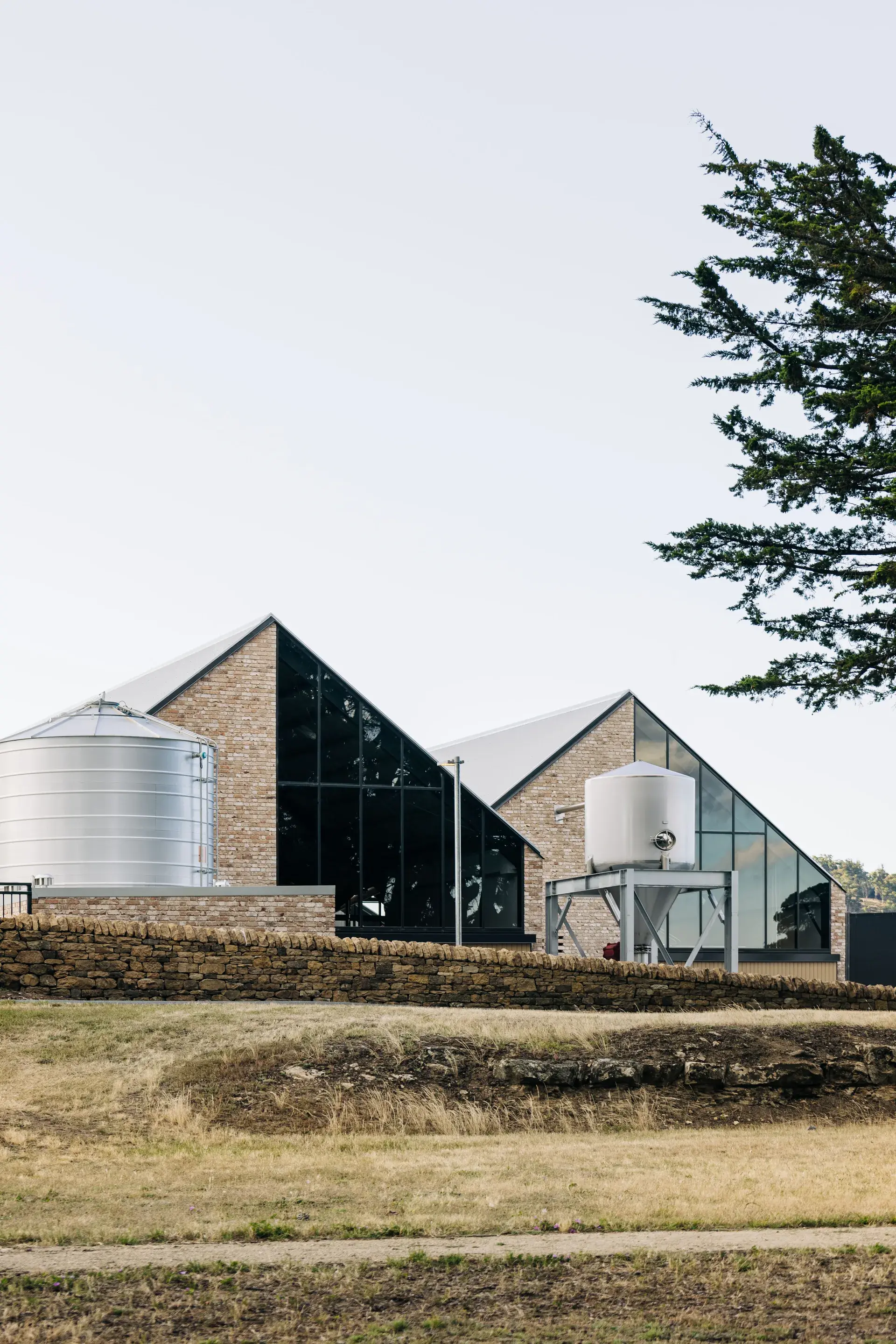 Callington Mill Distillery buildings in Oatlands, Tasmania, showcasing sharp gabled roofs of pale brick and glass alongside industrial silos, elevated behind an original historic stone wall to blend contemporary industry with heritage landscape.