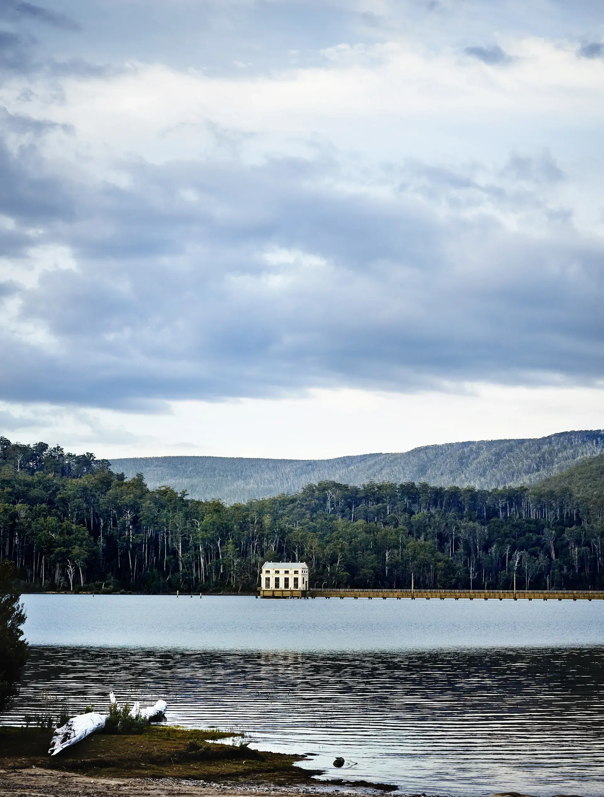 A wide shot of a historic white pump house building at the end of a long pier on a lake, surrounded by a dense green forest and distant hills.