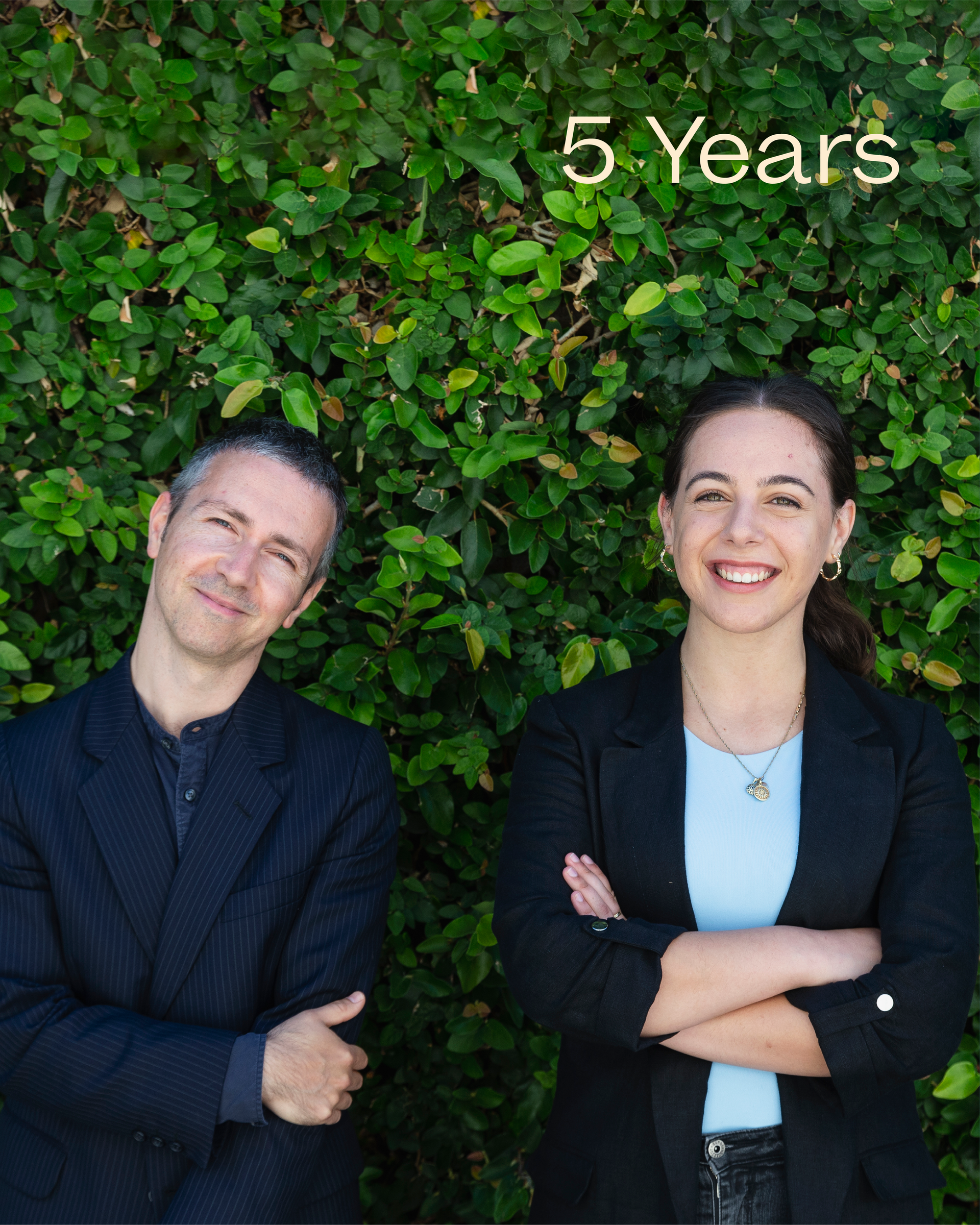 Jet O'Rourke and Anastasia Mavrides standing relaxed side by side, smiling against a backdrop of a lush green hedge. Both team members are wearing formal clothes, and text reading '5 Years' is overlaid on top.