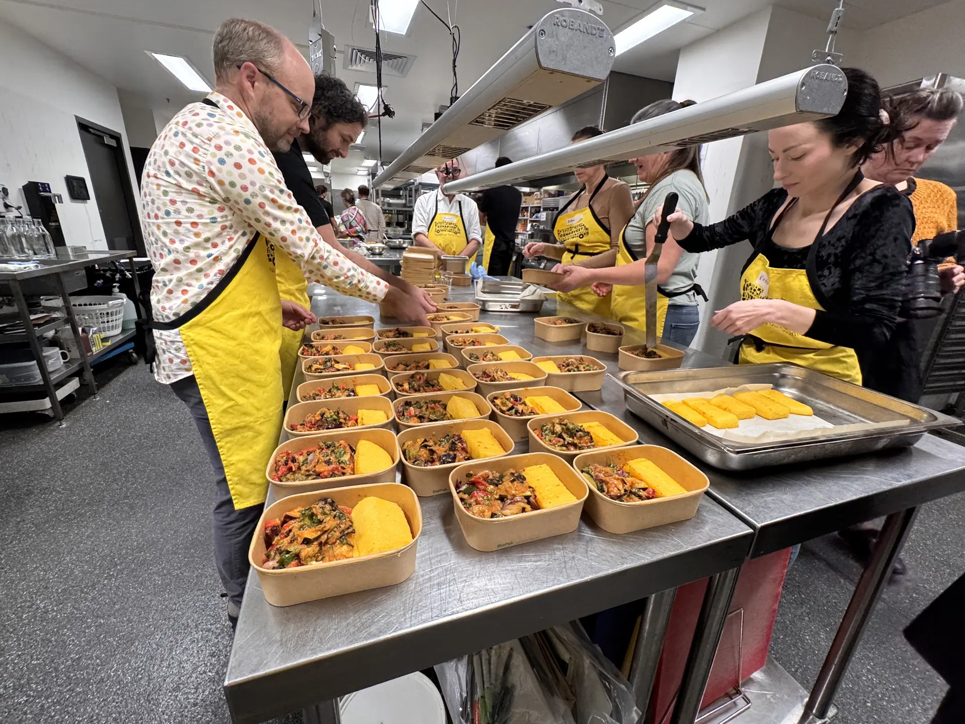 Cumulus Studio staff wearing yellow OzHarvest aprons work together to plate and package meals during a Cooking for a Cause workshop in Hobart, Tasmania.