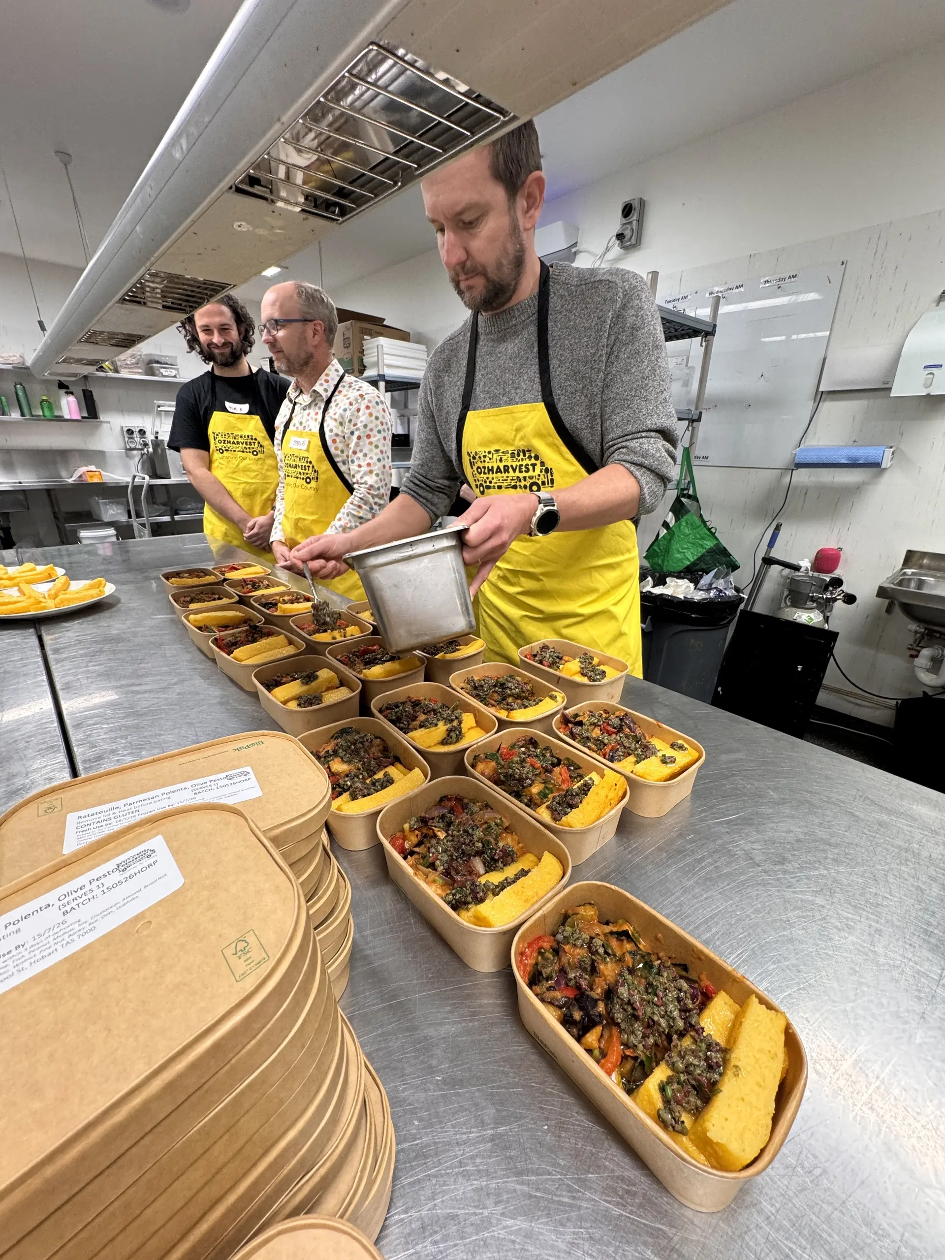 Cumulus Studio staff in yellow OzHarvest aprons add the finishing touches to rows of freshly prepared meals, ready for delivery to local people in need, during a Cooking for a Cause workshop in Hobart, Tasmania.