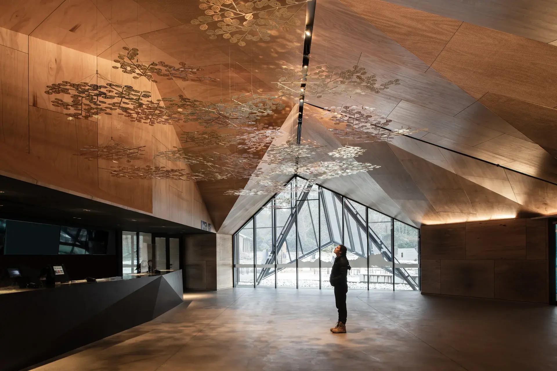 The dramatic interior of the Cradle Mountain Visitor Centre, featuring a soaring, faceted plywood ceiling that mimics the surrounding mountain peaks. A visitor stands in the minimalist lobby, looking up at a delicate floating art installation suspended beneath a large triangular window.