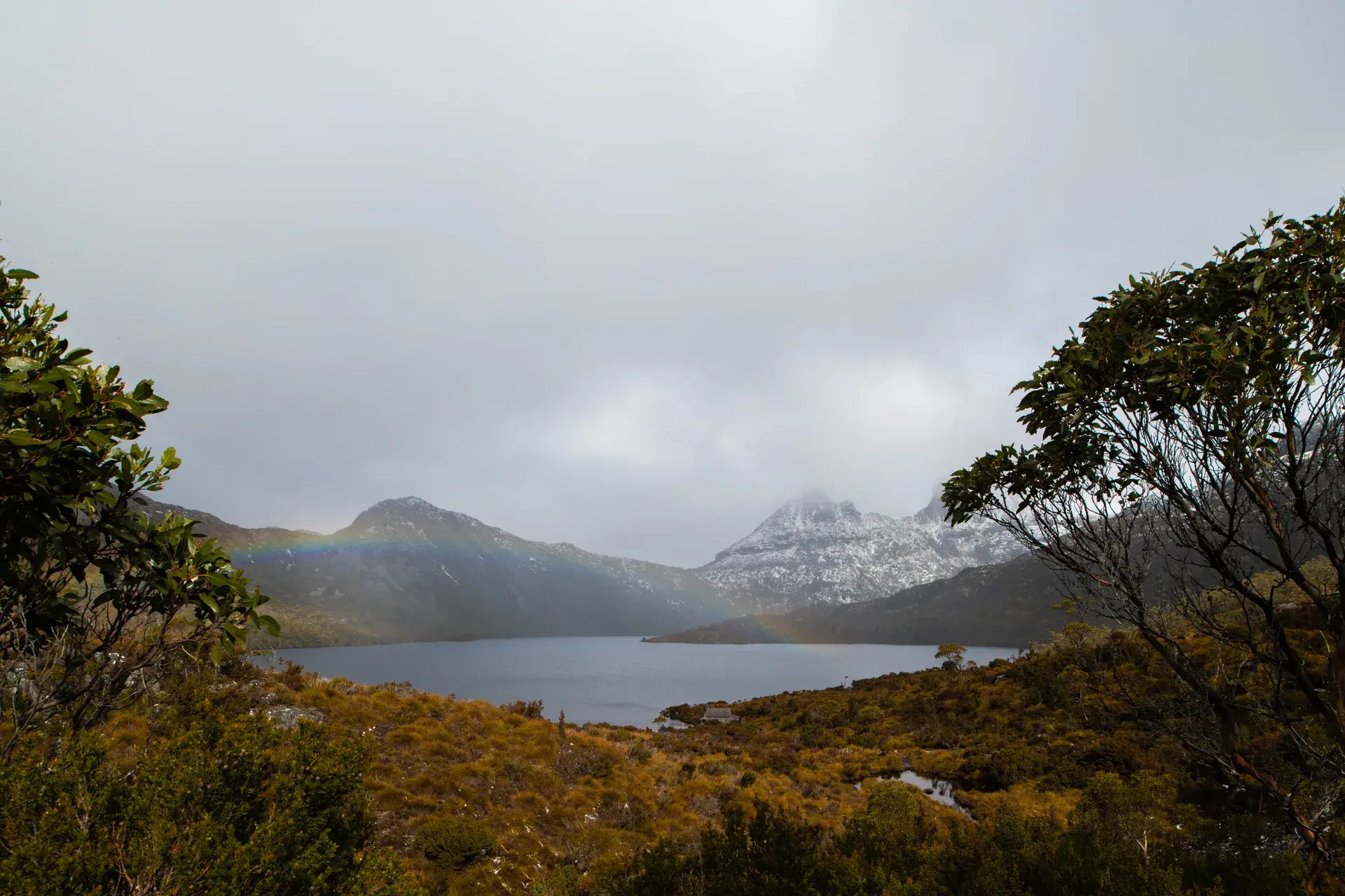 A wide shot of Dove Lake at the base of Cradle Mountain, Tasmania, with snow-dusted jagged peaks rising behind the water and a faint rainbow appearing under a heavy, overcast sky.