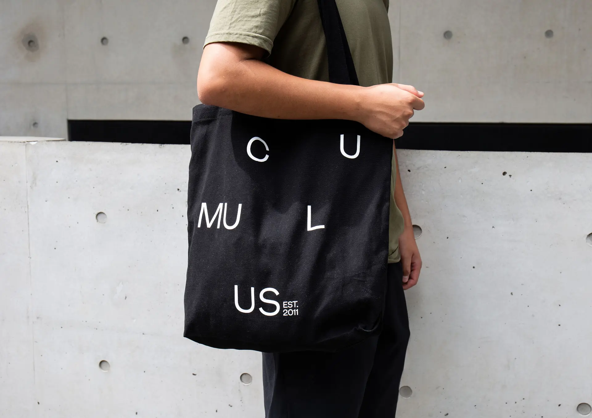 A photograph of a person holding a black tote bag featuring Cumulus’ new adaptive typographic identity by For the People, with dispersed white lettering forming “CUMULUS” and “EST. 2011” across the surface.