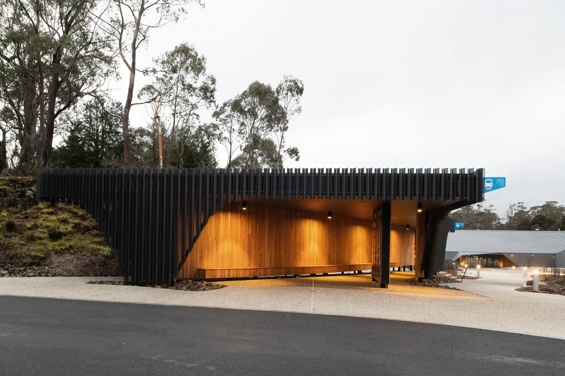 At Cradle Mountain Visitors Centre, a striking black slatted structure at dusk features a warm, timber-lined alcove with integrated seating, creating a glowing focal point against the darkening trees and overcast sky.
