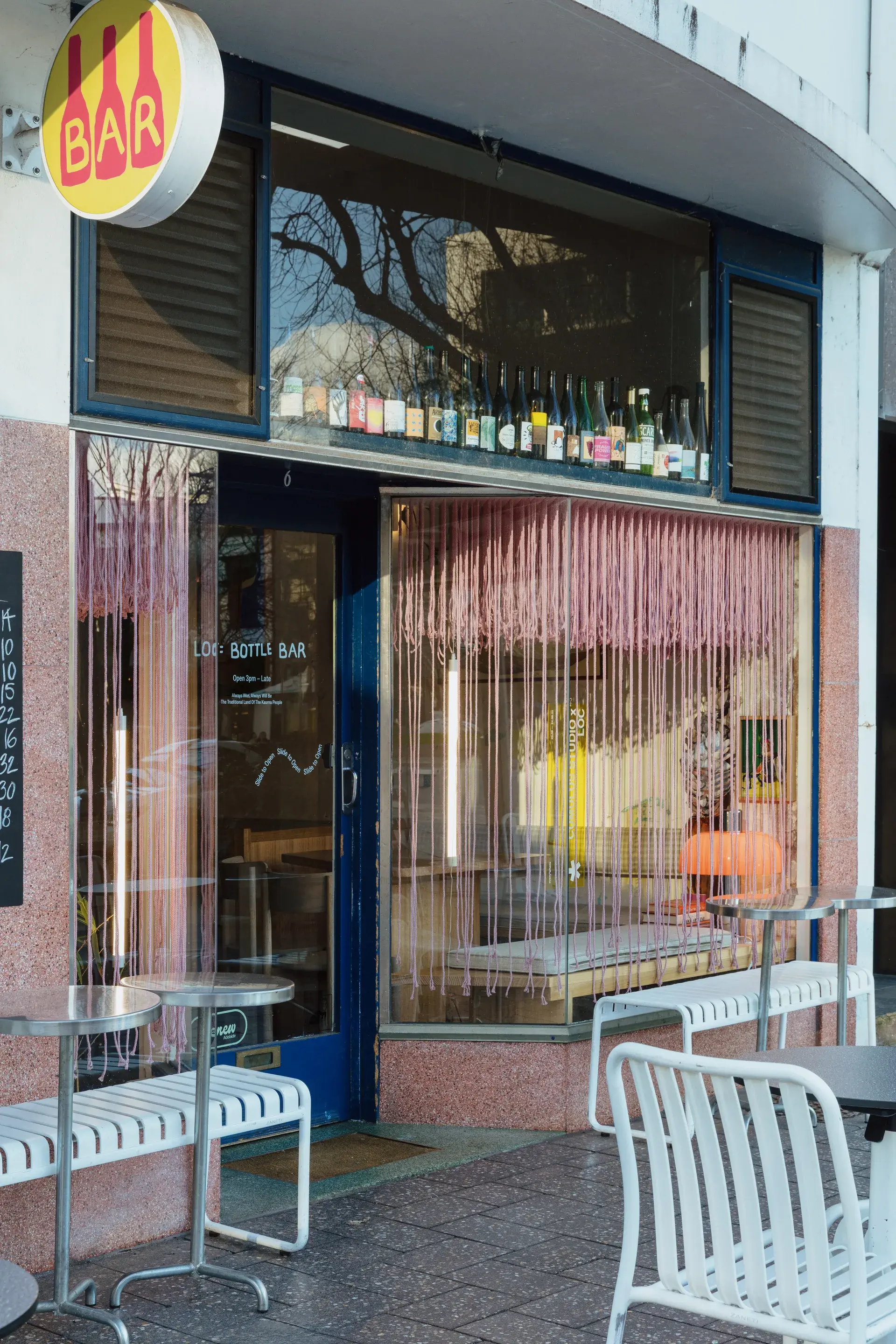 The storefront of "LOC BOTTLE BAR" featuring a blue door, a pink rope ceiling installation in the window, and outdoor seating with metal tables and white benches.