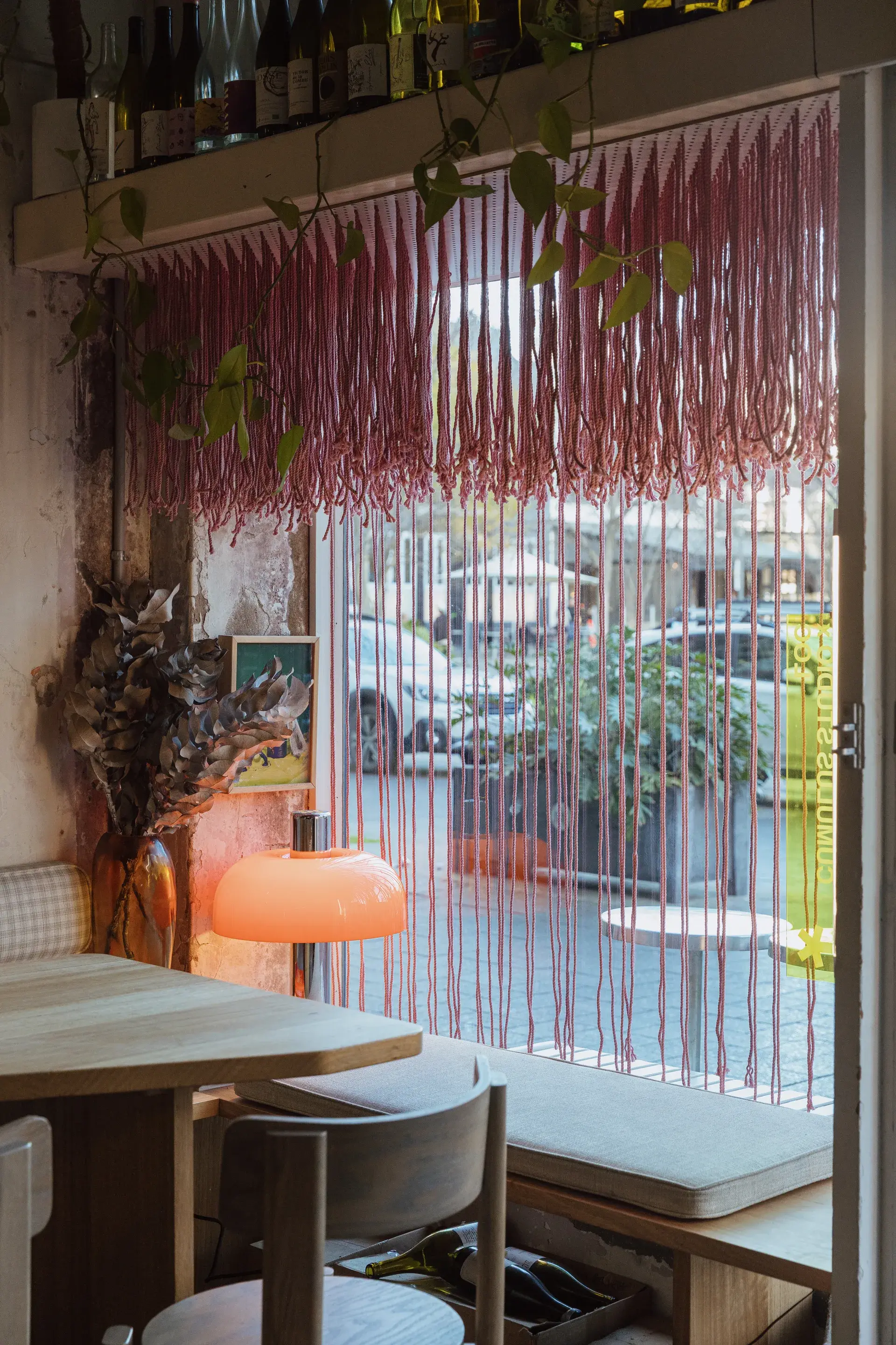 An interior view of a cozy wine bar seating area featuring a wooden table, a glowing orange mushroom lamp, and a pink rope curtain looking out onto a street.