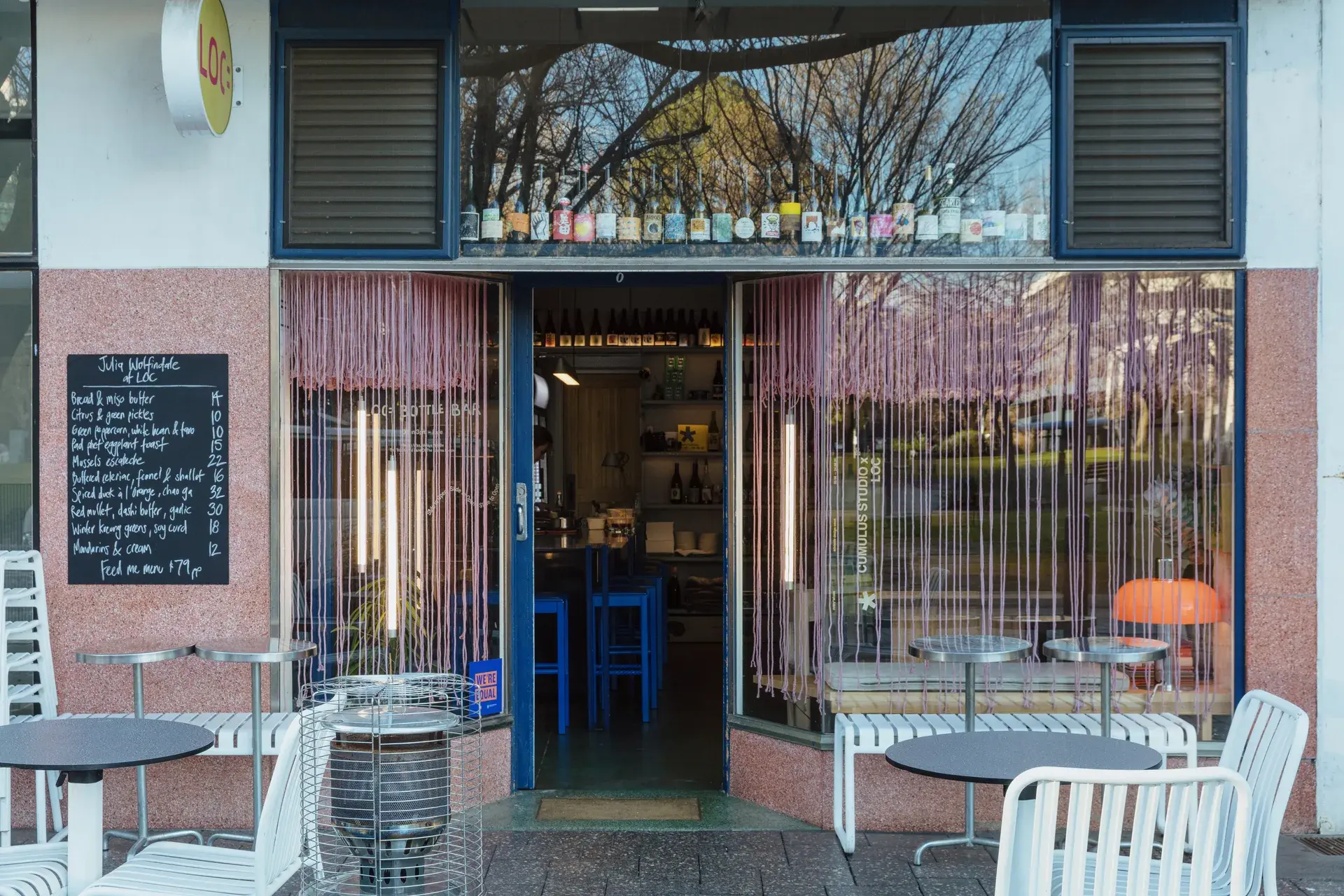 Wide storefront view of the wine bar with blue-framed windows, outdoor bistro seating, and a handwritten chalkboard menu displaying daily specials.