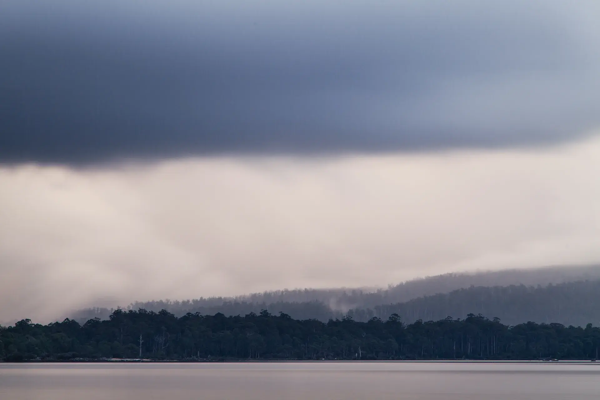 A moody, atmospheric landscape of a calm Tasmanian waterway reflecting low-hanging dark storm clouds and mist-covered forested hills in the distance.
