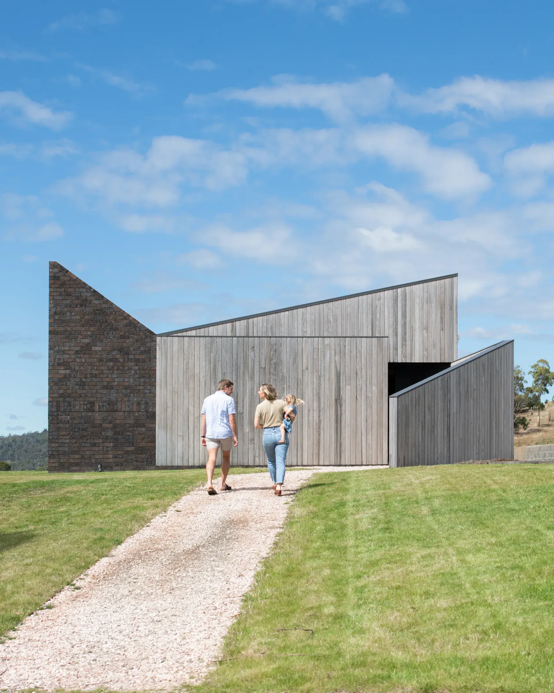 A view of a building with a sharp, angular silhouette made of silver-weathered timber and dark stone, featuring a family walking up a gravel path toward the minimalist entrance set against a vast rural landscape.