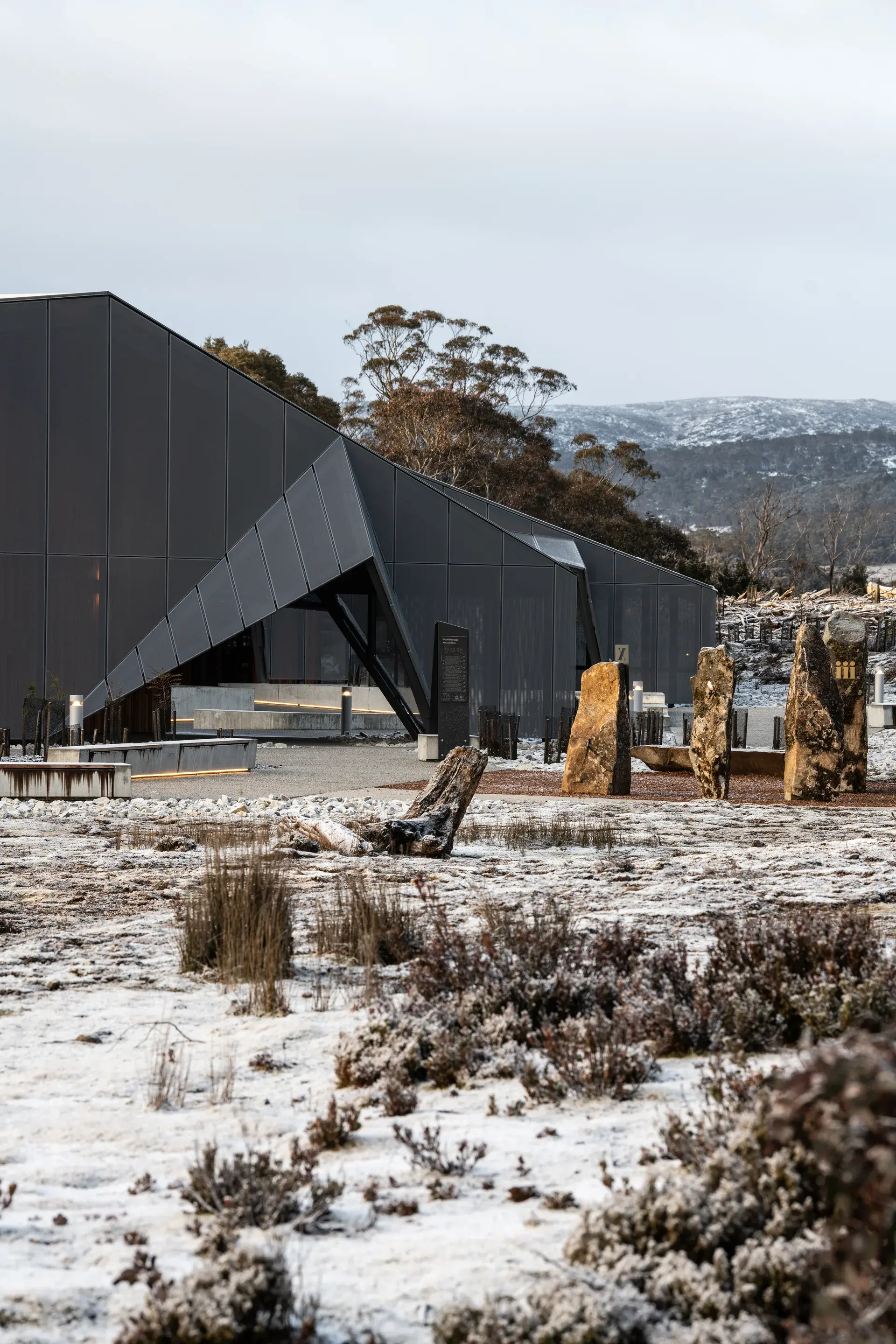 The Cradle Mountain Visitor Centre, featuring dark, sharp-angled modern facades and triangular entryways that mimic the rugged peaks of the surrounding alpine landscape, set against a backdrop of snow-dusted heath and jagged dolerite-style monoliths.