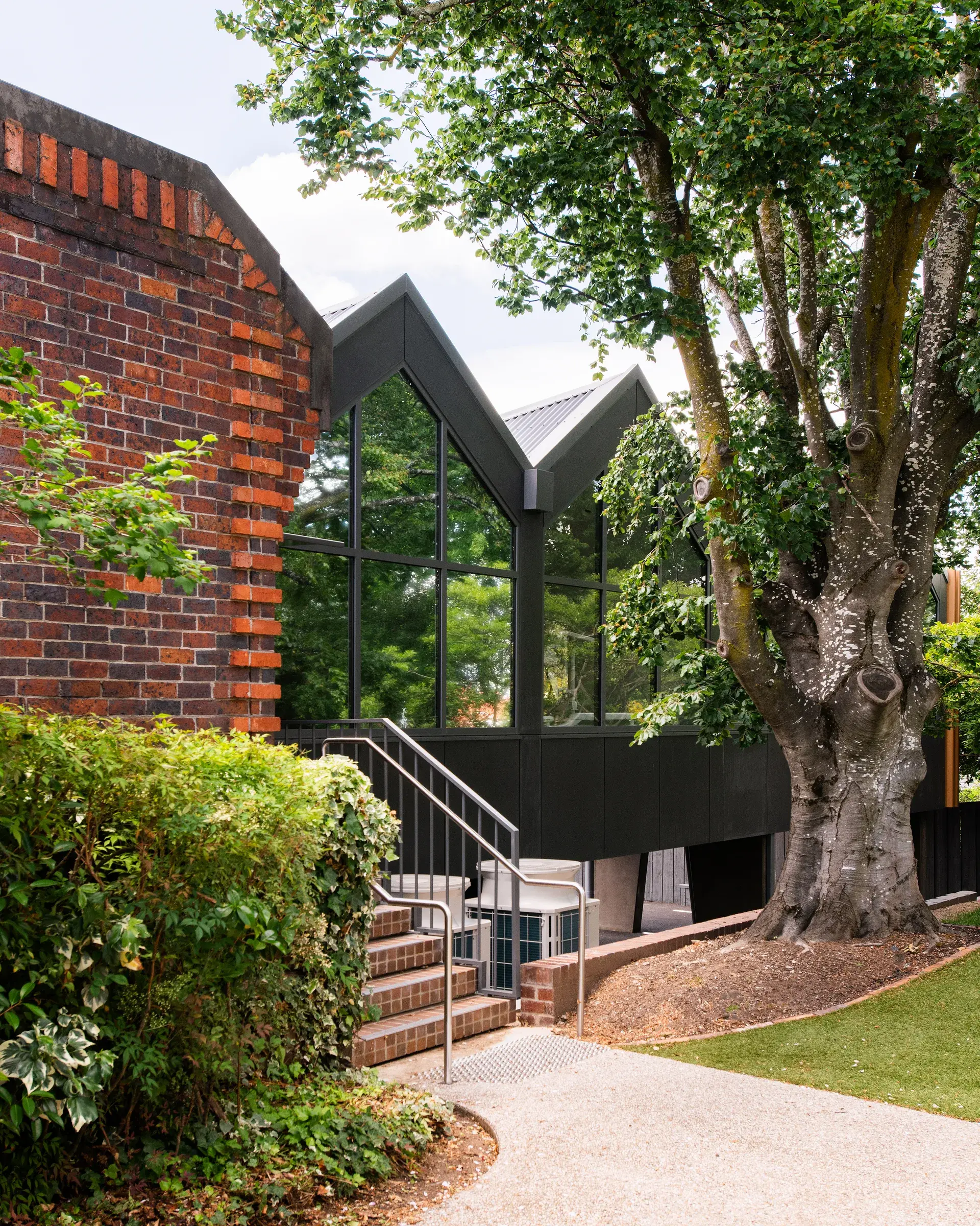 Exterior view of a modern building with large gabled glass windows and dark metal siding, situated next to a traditional brick wall and a large, established tree.