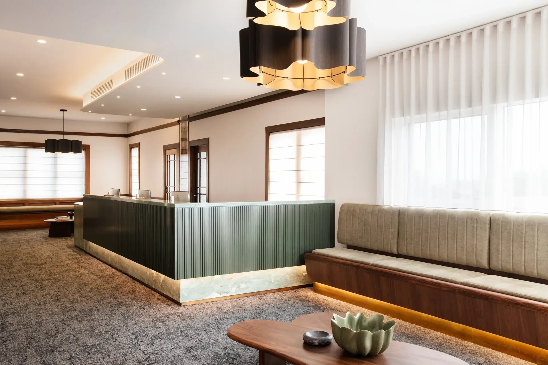 An elegant office lobby featuring a fluted dark green reception desk with under-lighting, sleek wooden benches, and modern floral-shaped chandeliers.