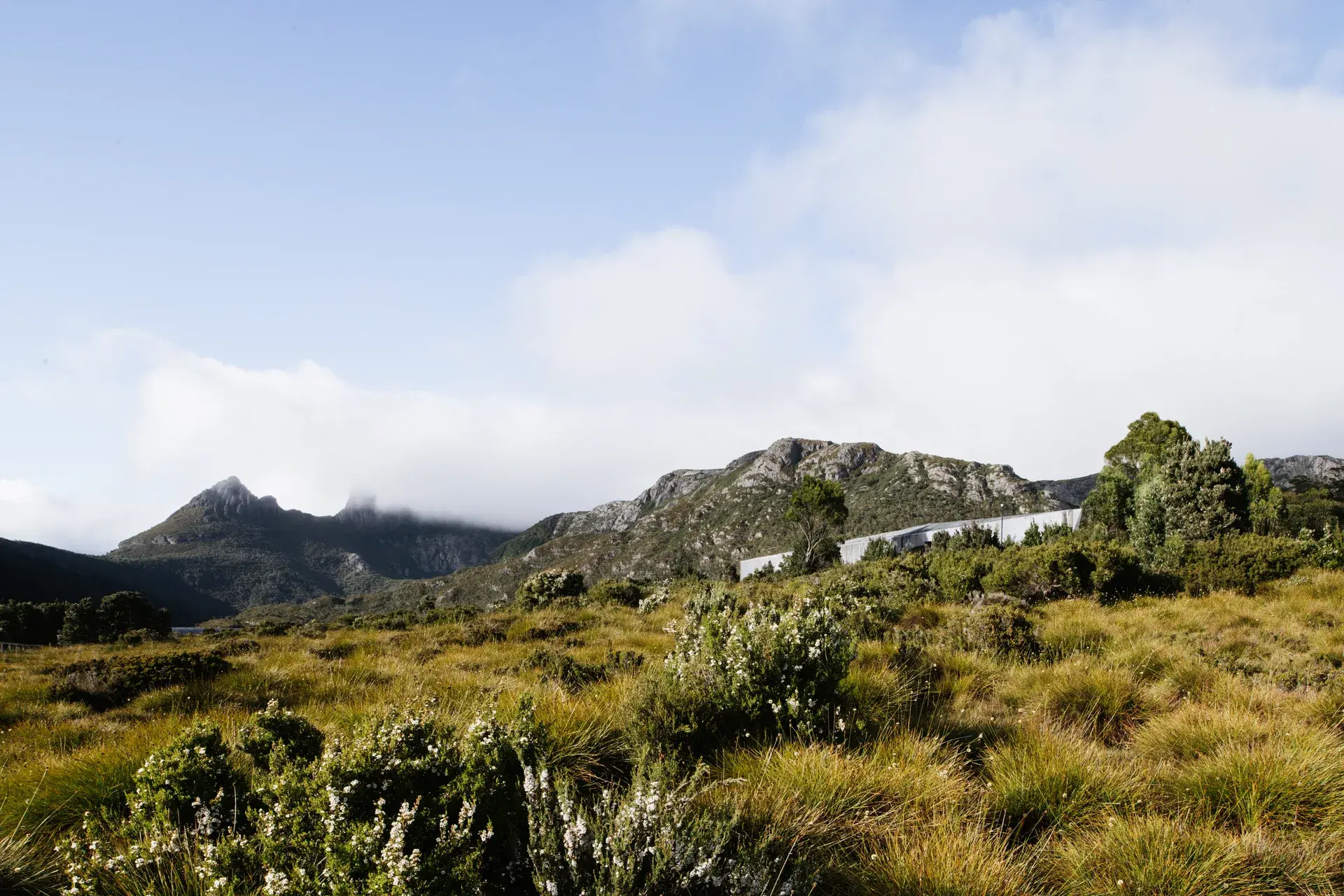 A long, shrouded building is tucked into a vast alpine landscape of rolling grassy plains and dense shrubbery, set against a backdrop of mist-covered mountain peaks.