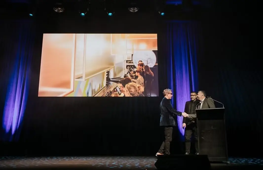 Cumulus Studio team members on stage accepting an award for Kosaten Launceston, shaking hands beside a podium with a large projected image of the restaurant interior behind them.