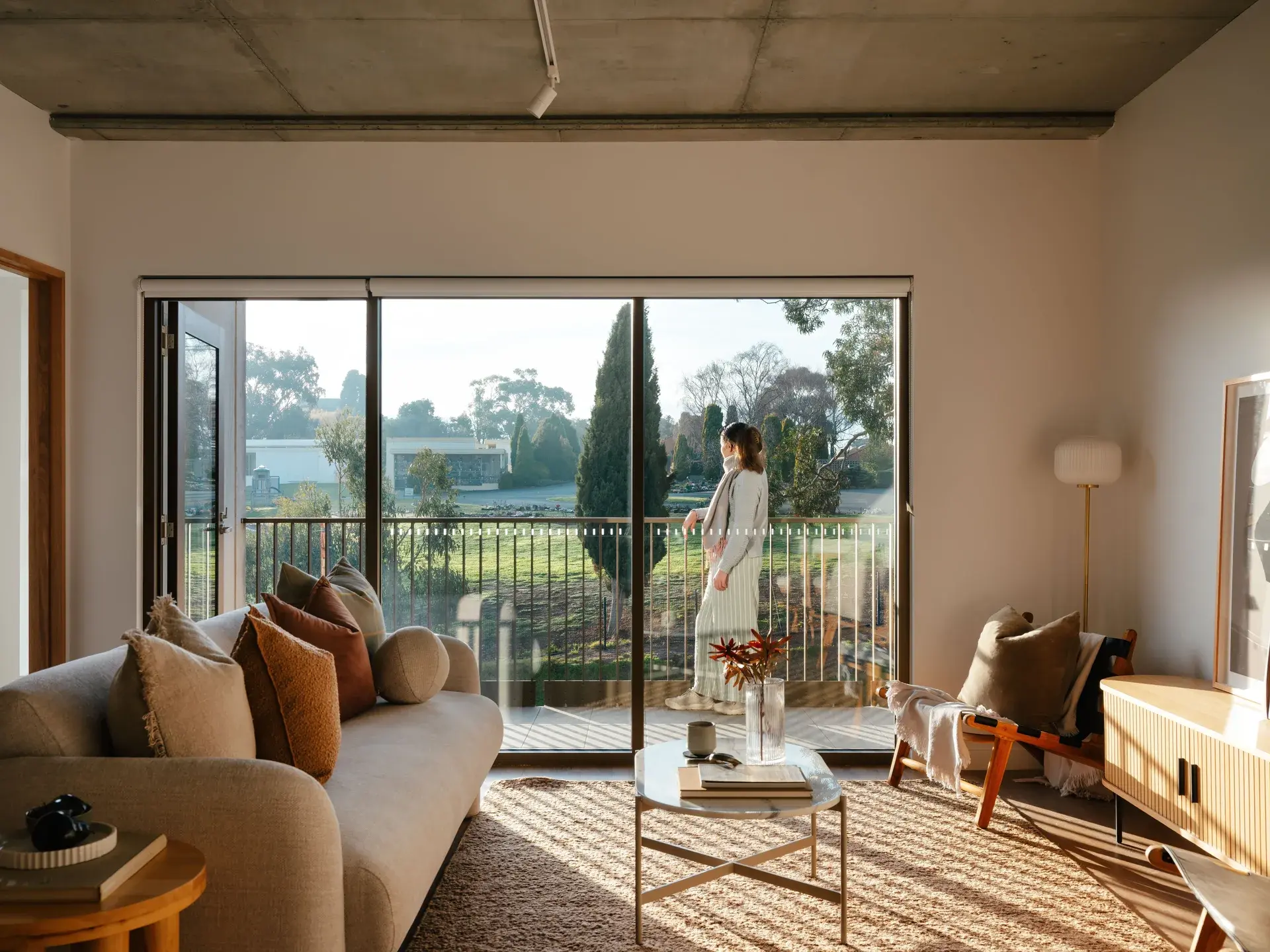 A person on their balcony, in the foreground is a lounge room with subtle light coming through.