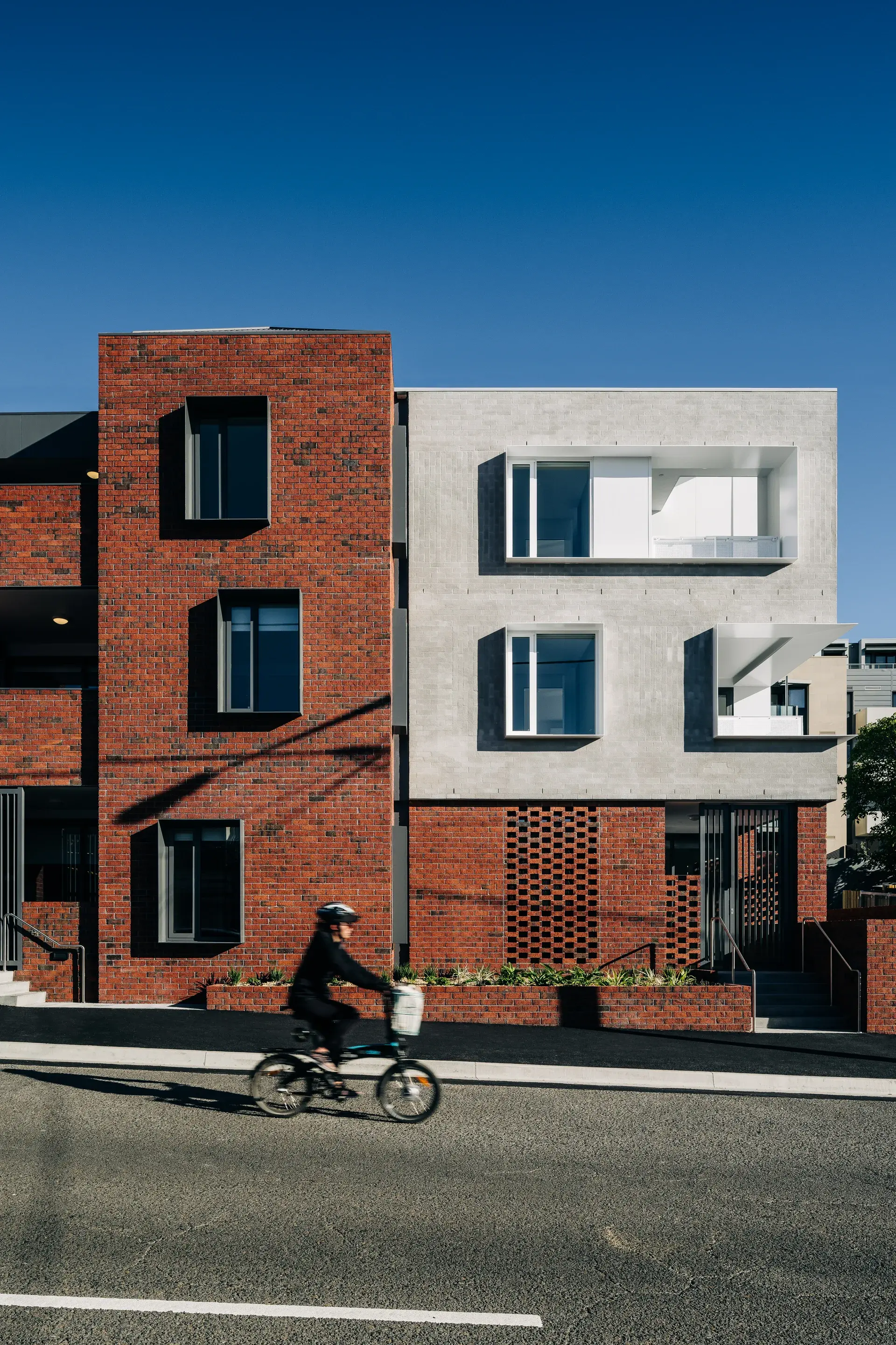 A red brick base anchors the building to the street, with lighter masonry volumes above featuring deep-set windows and projecting balconies. Varied brick textures and recessed openings create a layered façade, while a cyclist passes in the foreground.