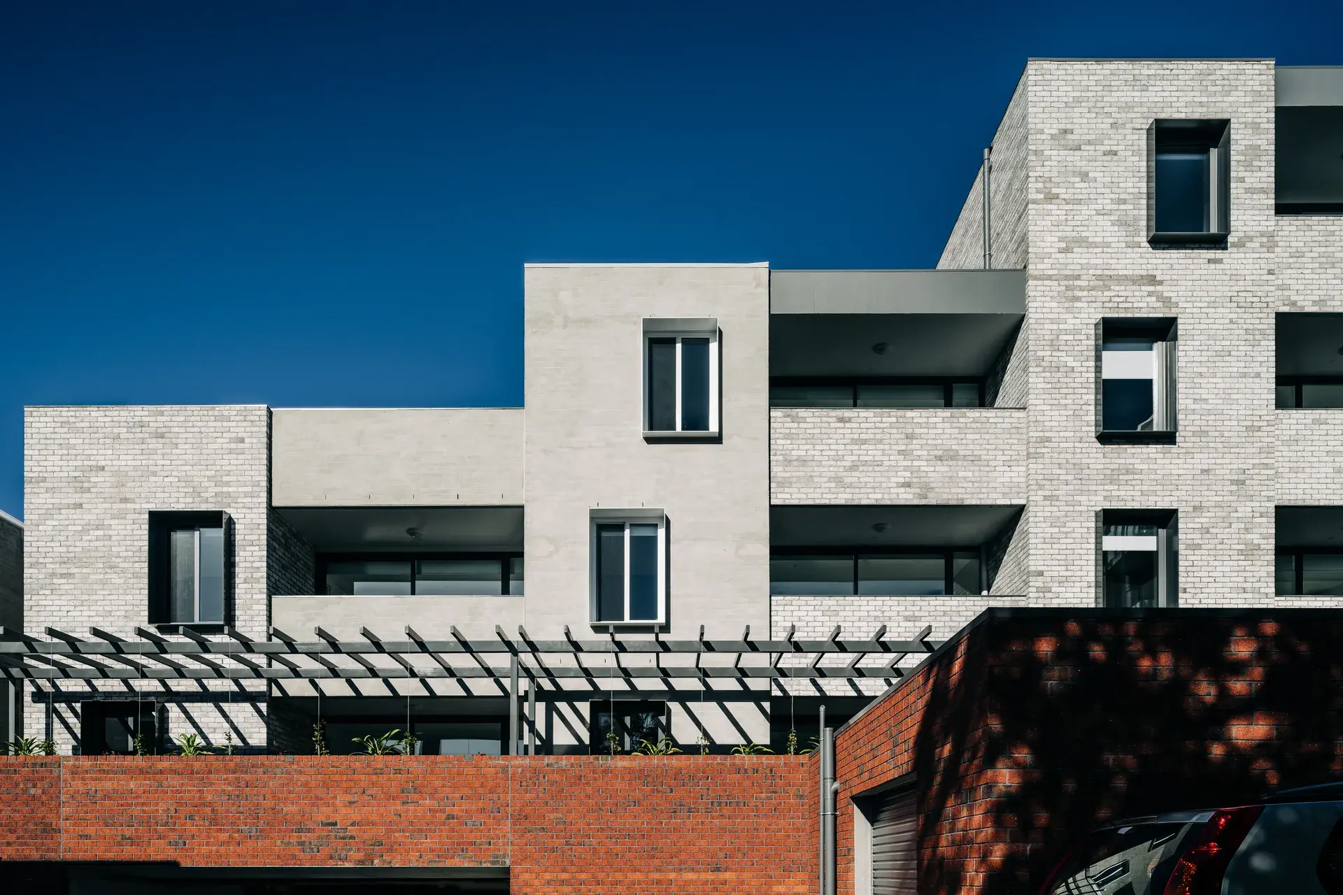 A series of stepped, light-coloured masonry volumes rise above a red brick podium, with recessed balconies and deep window reveals. A black steel pergola extends along the lower level, shading planter boxes filled with greenery and reinforcing the building’s horizontal rhythm.