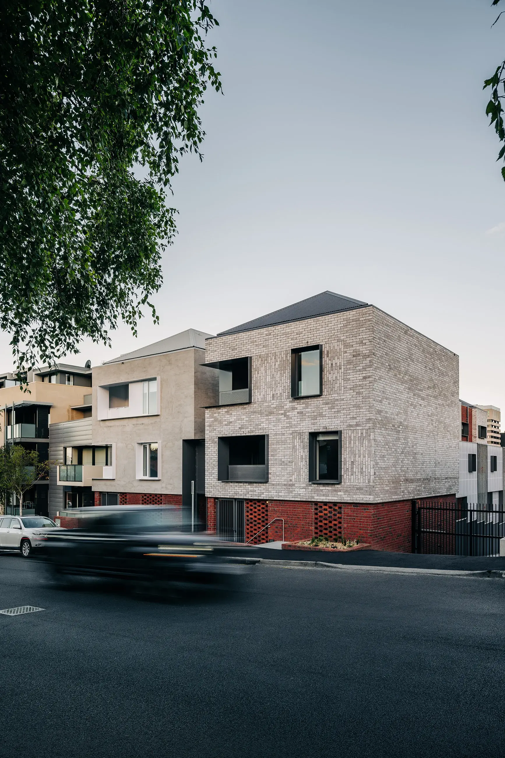 Two modern multi-story buildings both feature a consistent lower strip of red brick, with one finished in a pale brick above and the other a pale render. Both have a pyramid hip roof and deep-set windows standing along a busy street lined with parked cars.