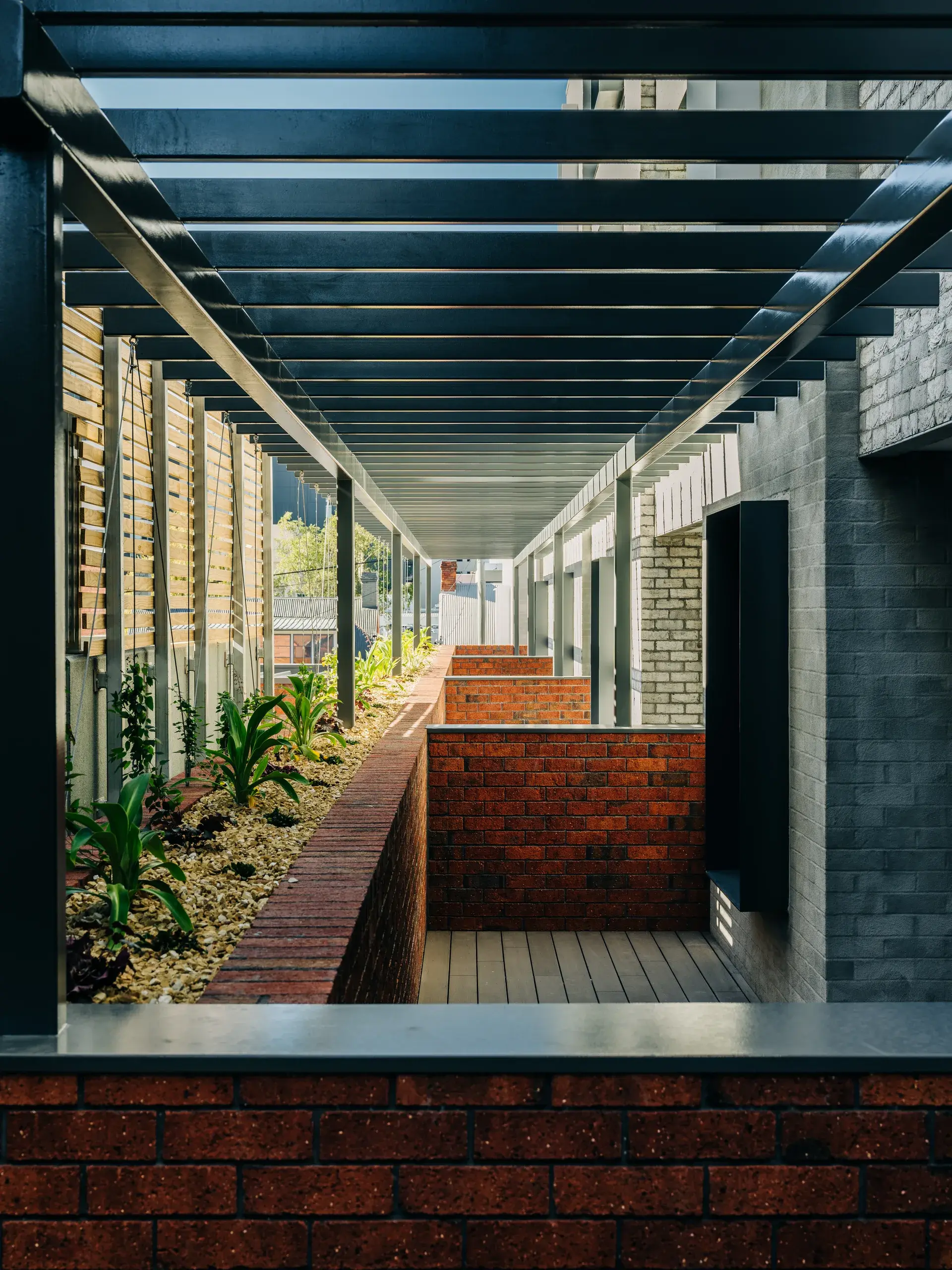 A long line of balconies are covered by a black steel pergola and features brick planter boxes filled with green plants and gravel.