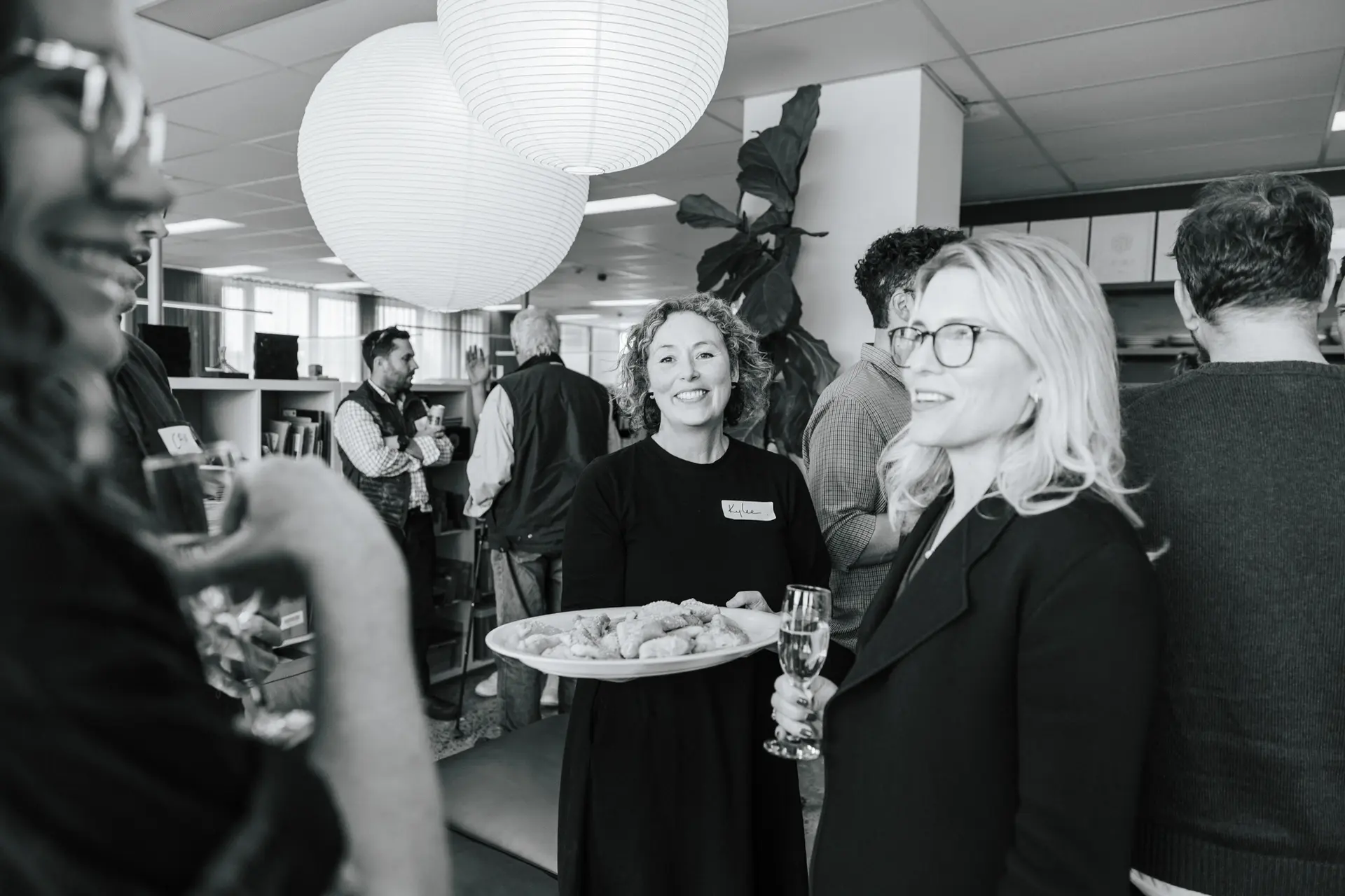 Black and white photo of Kylee smiling holding a platter of appetisers at a social gathering in the Hobart office with paper lantern lights above.