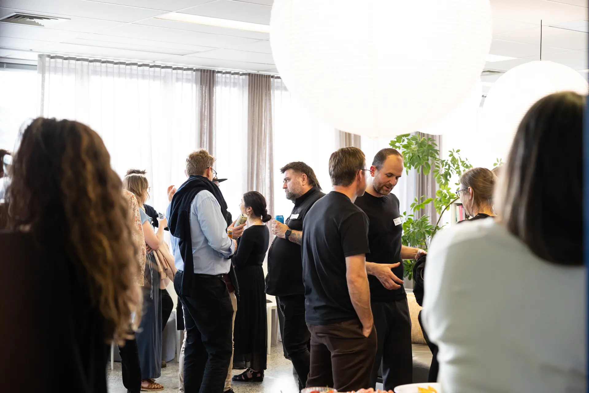 A brightly lit social gathering in a modern office space, with groups of people talking under large, white spherical pendant lights.