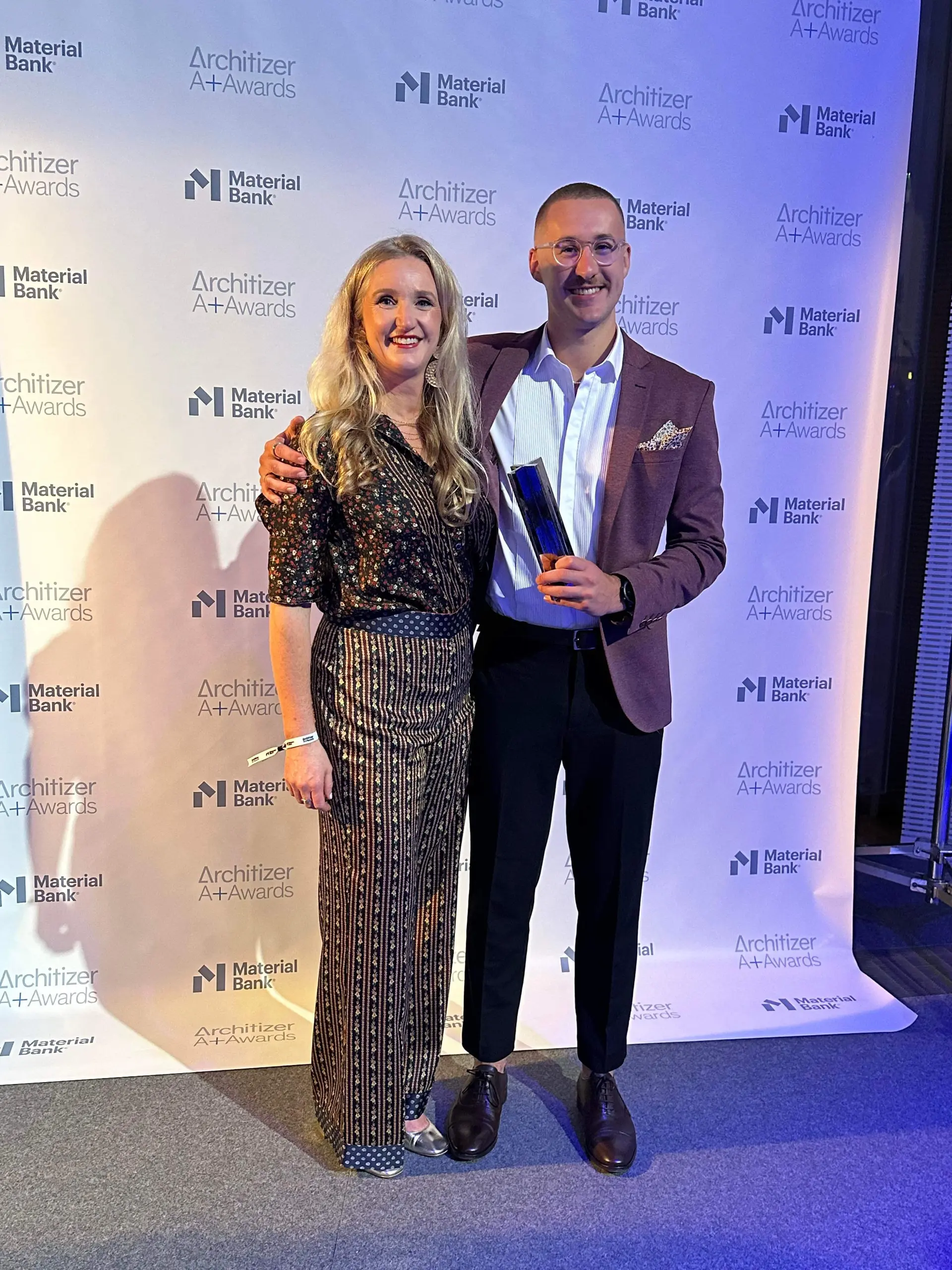 Jenn Heggarty and Jarvis Heggarty-Henderson smiling, hold the Architizer A+ Awards on behalf of Cumulus at the awards ceremony in Paris. Jen is wearing a patterned floral blouse and pants, and Jarvis wears a suit with purple blazer.
