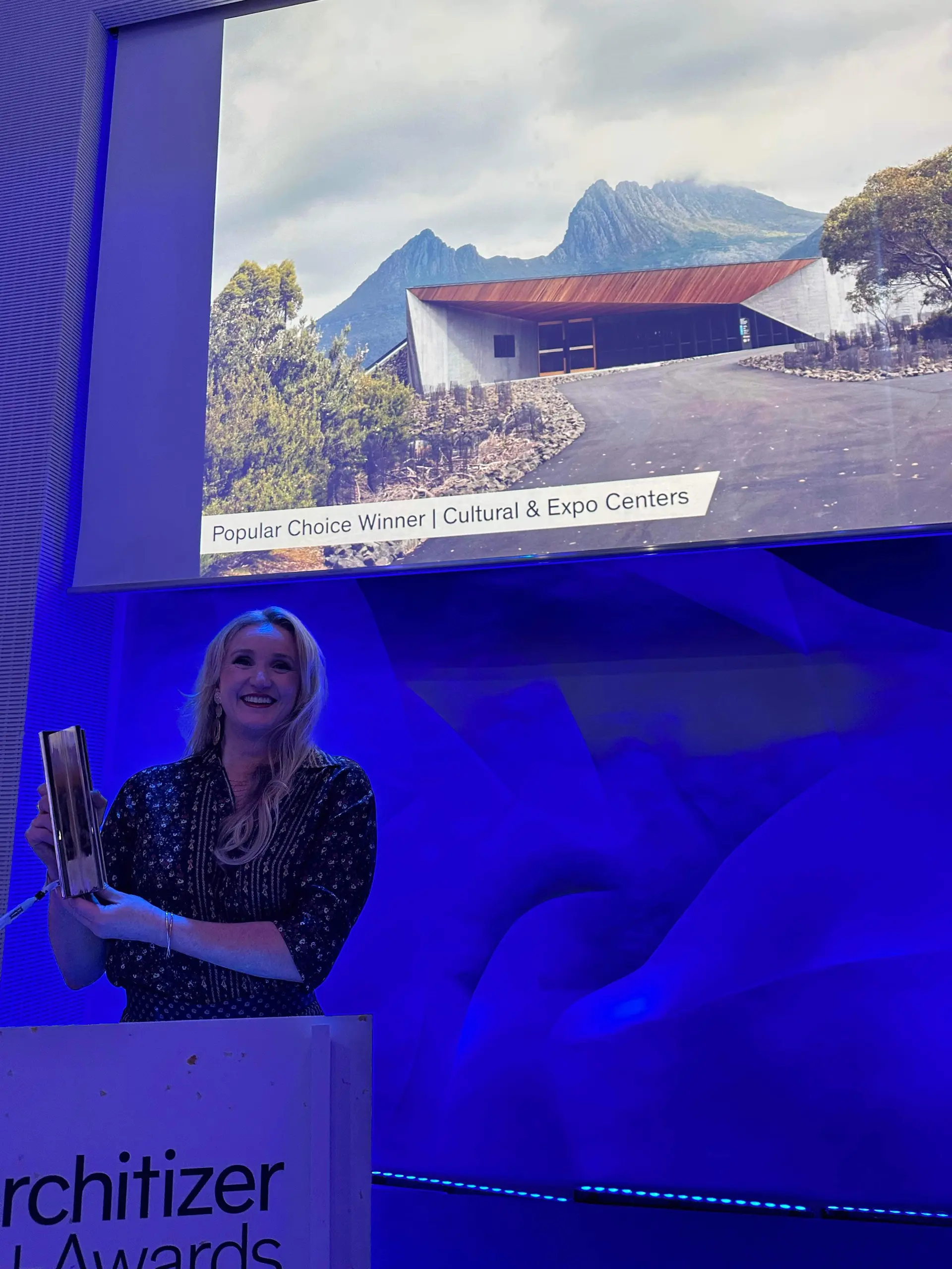 Jenn Heggarty accepts the Popular Choice Winner award in the Cultural & Expo Center category for Dove Lake Viewing Shelter, smiling and holding the award at a podium while the building is projected on the wall behind her.