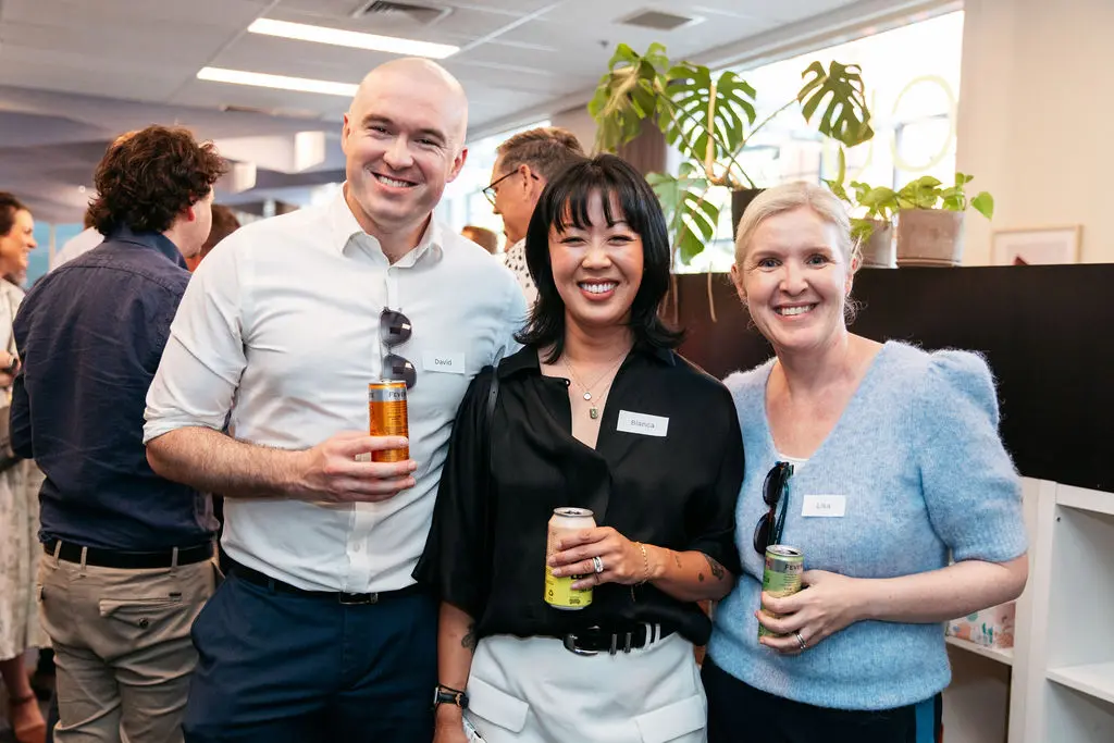 Three smiling people holding drinks and wearing name tags while posing together at a brightly lit social event with indoor plants in the background.
