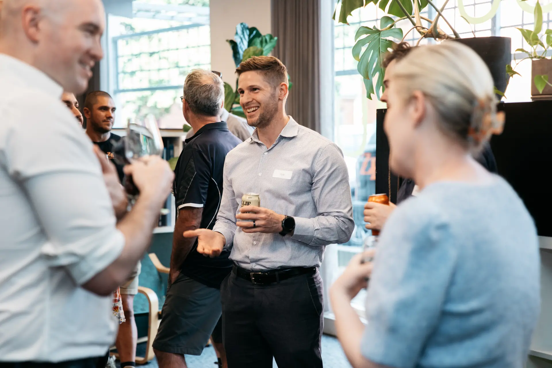 A smiling man holds a drink and chats with others at a bright indoor social event.