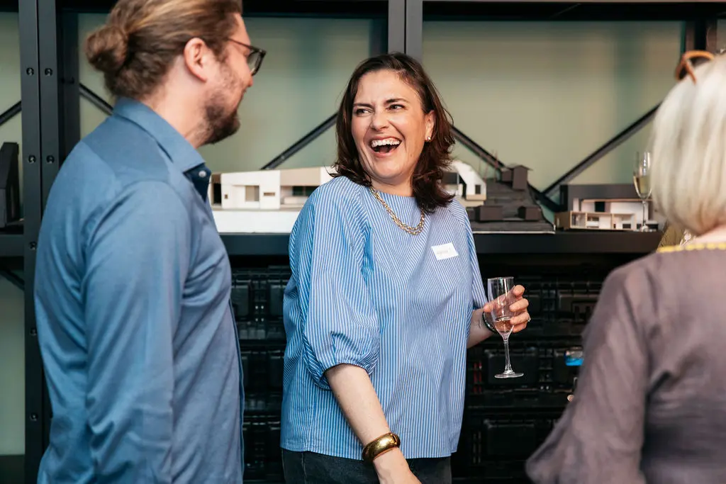 A woman holding a drink and talking with colleagues at a social event, with architectural models on a shelf in the background.