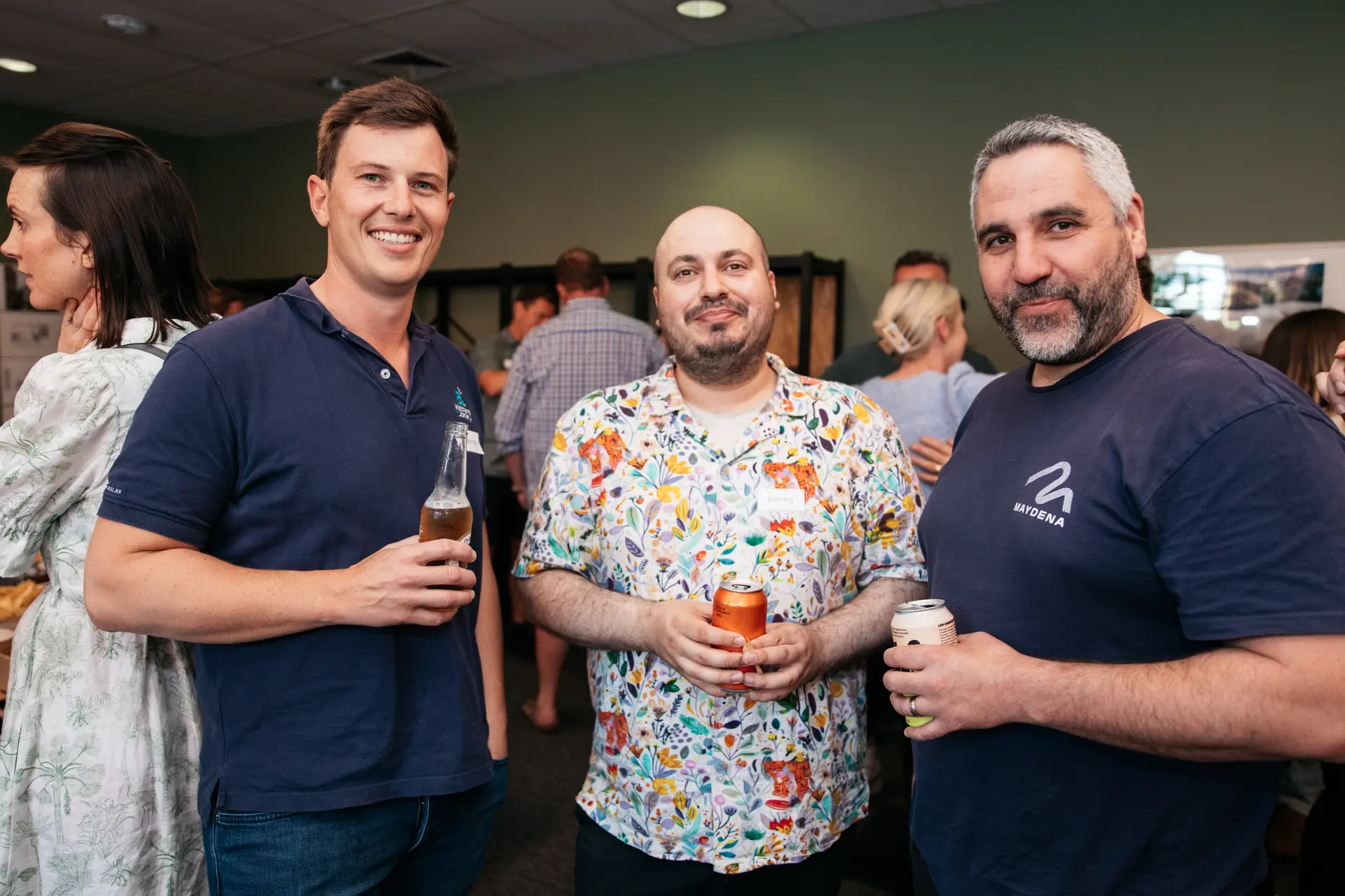 Three men holding drinks and smiling at an indoor social gathering, with one man wearing a vibrant floral patterned shirt.