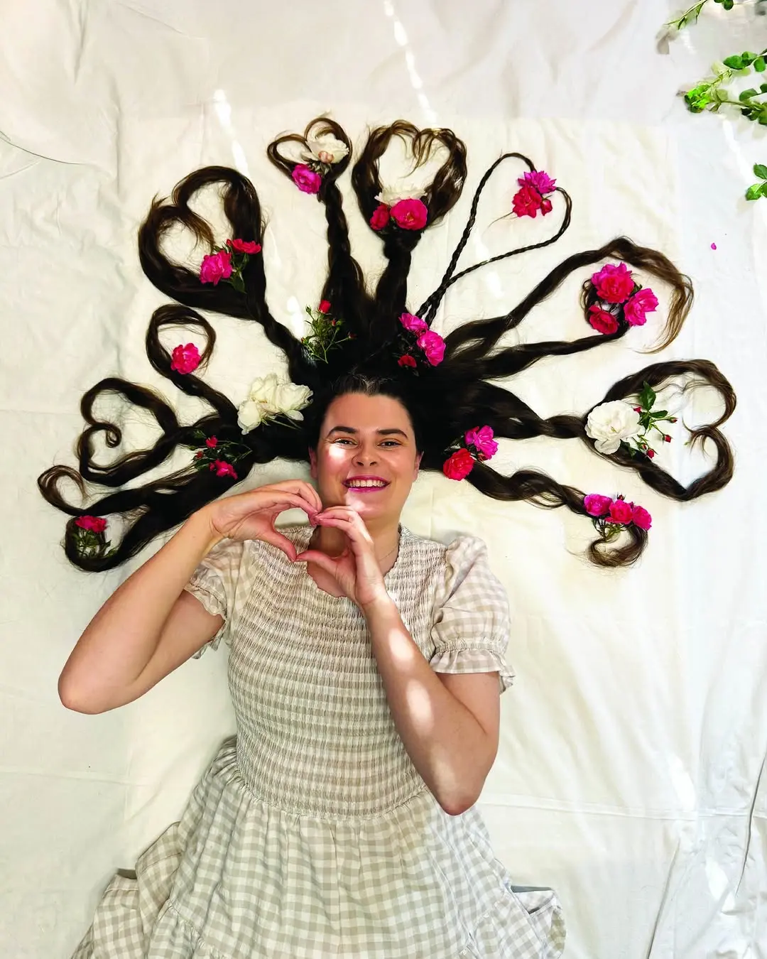 Maddie lies down, her long dark hair spread out and styled into several heart shapes adorned with pink and white roses. She is smiling and making a heart shape with her hands.
