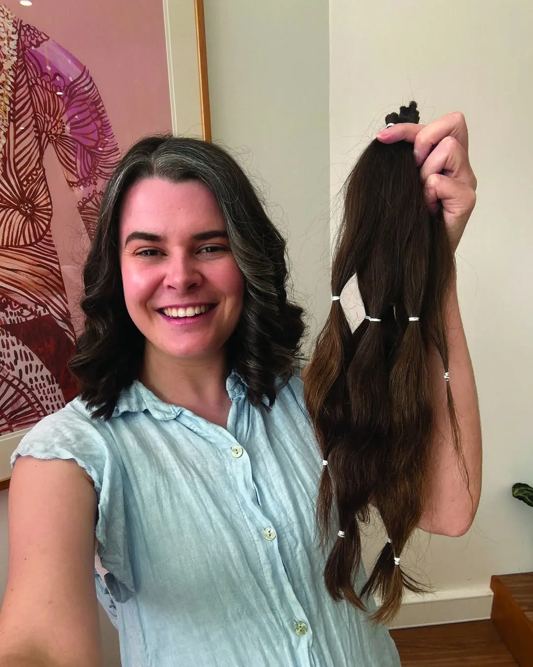 Maddie smiles with shoulder-length wavy hair holds up her long, thick ponytails of dark brown hair that has been cut off and sectioned with white elastics for donation.