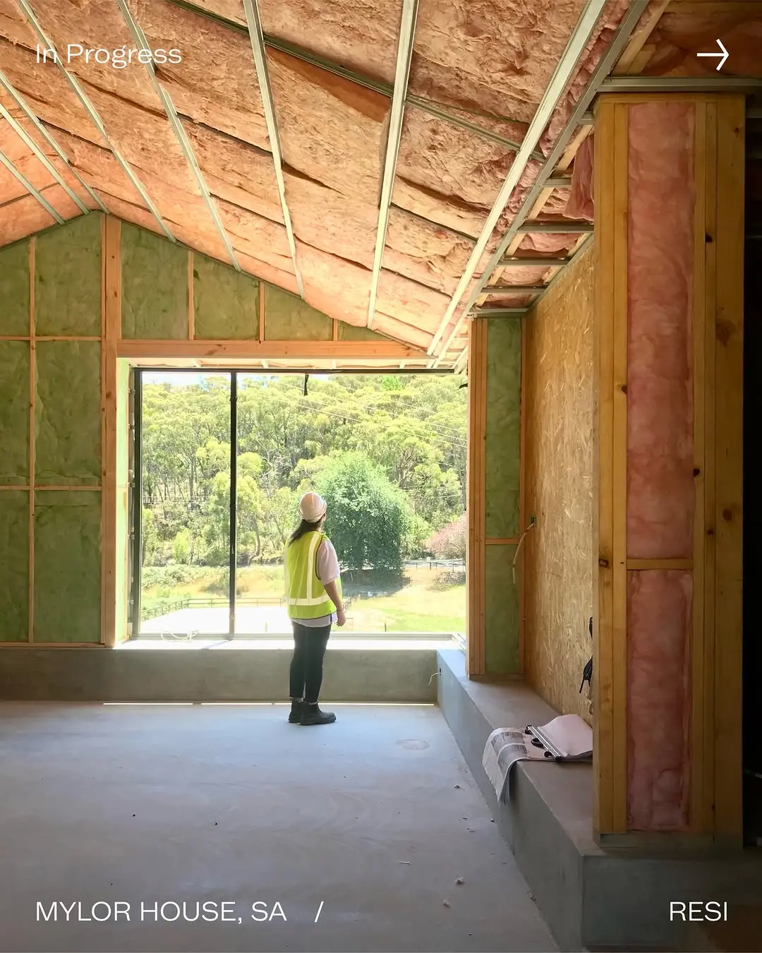 The Mylor House under construction, with a lady looking out a large window at a lush green landscape. The room shows exposed timber framing and pink and green insulation.