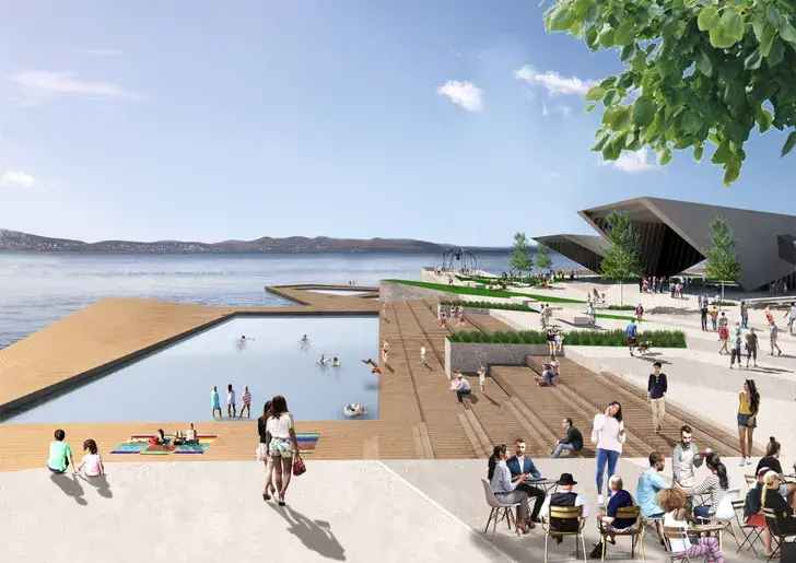 Waterfront scene featuring a large tidal swimming pool integrated into a timber boardwalk, surrounded by terraced seating, cafés, and geometric pavilion buildings, with people swimming, sunbathing, and socialising along the edge of the harbour.