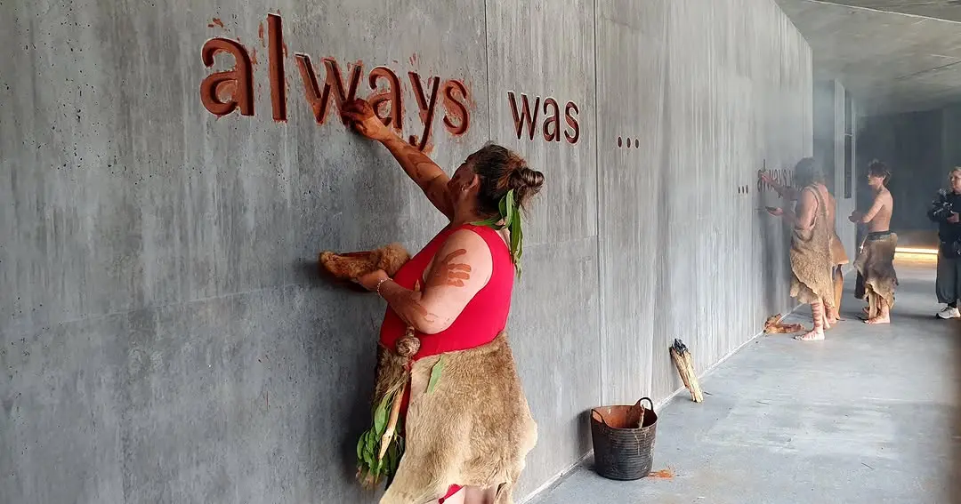 A Tasmanian Aboriginal woman uses red ochre to colour the carved letters of the phrase "always was..." on a concrete wall in Dove Lake Viewing Centre, performs a ritual that reclaims contemporary architectural space for the palawa people.