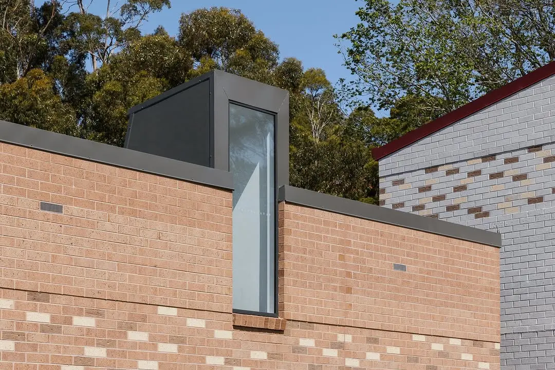 A close-up architectural detail of a modern tan brick building featuring a vertical glass window encased in a dark grey metal frame, with a light grey brick building and green trees in the background.