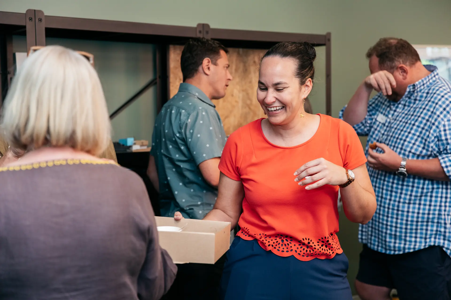 Anna with a large smile on her face is handing out catering in a cardboard box within a social gathering.