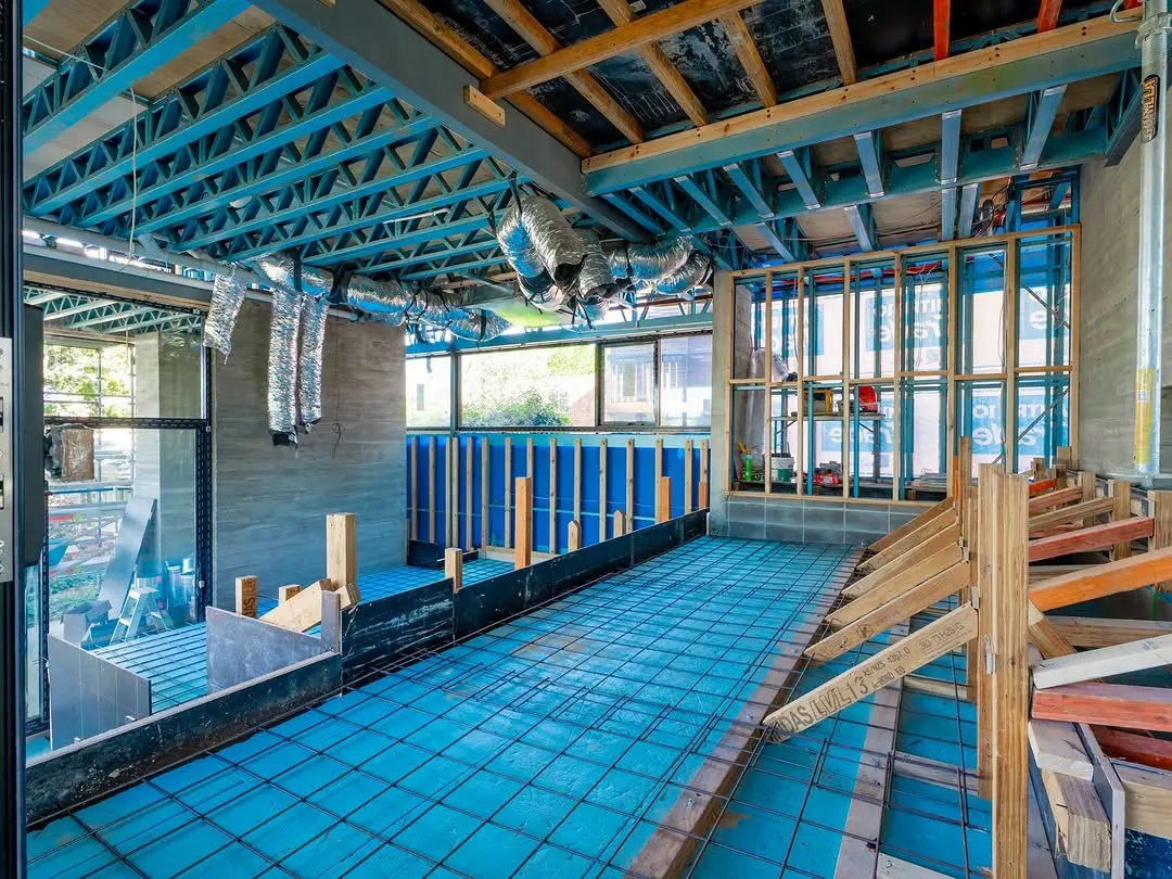 Interior view of the building under construction, showing a blue insulated floor with steel mesh, wooden wall framing, and exposed ceiling trusses with silver ventilation ducts.